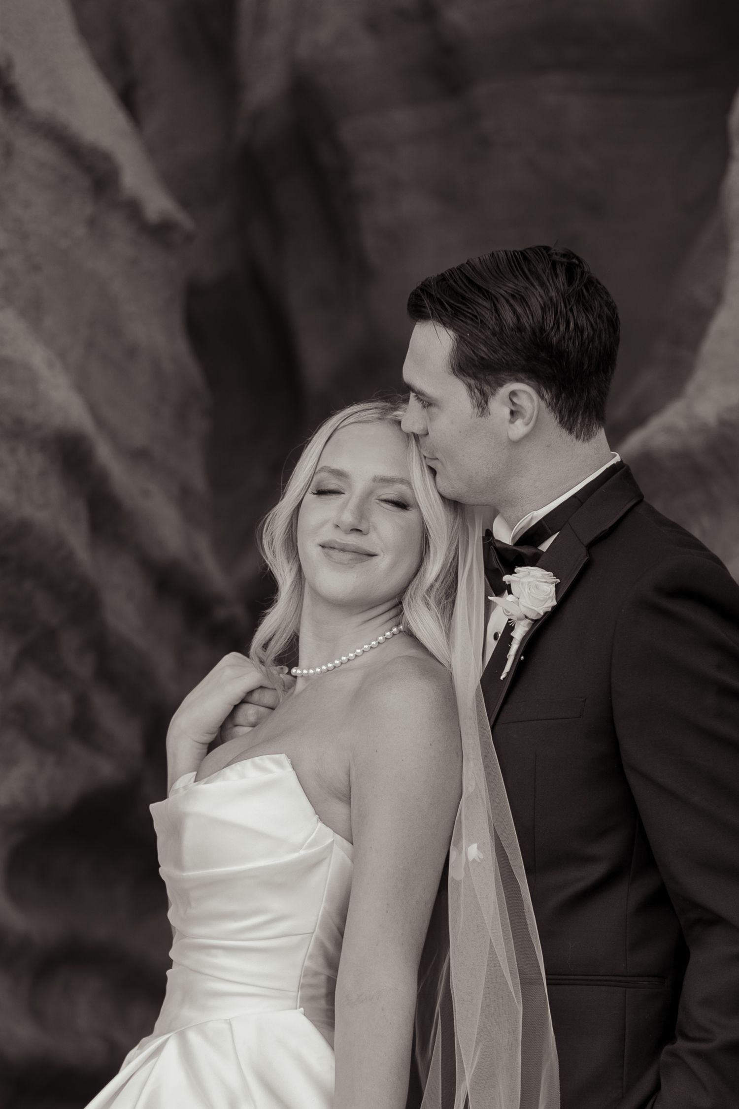 Black and white wedding photo of bride in strapless gown with pearl necklace as groom in tuxedo kisses her forehead.