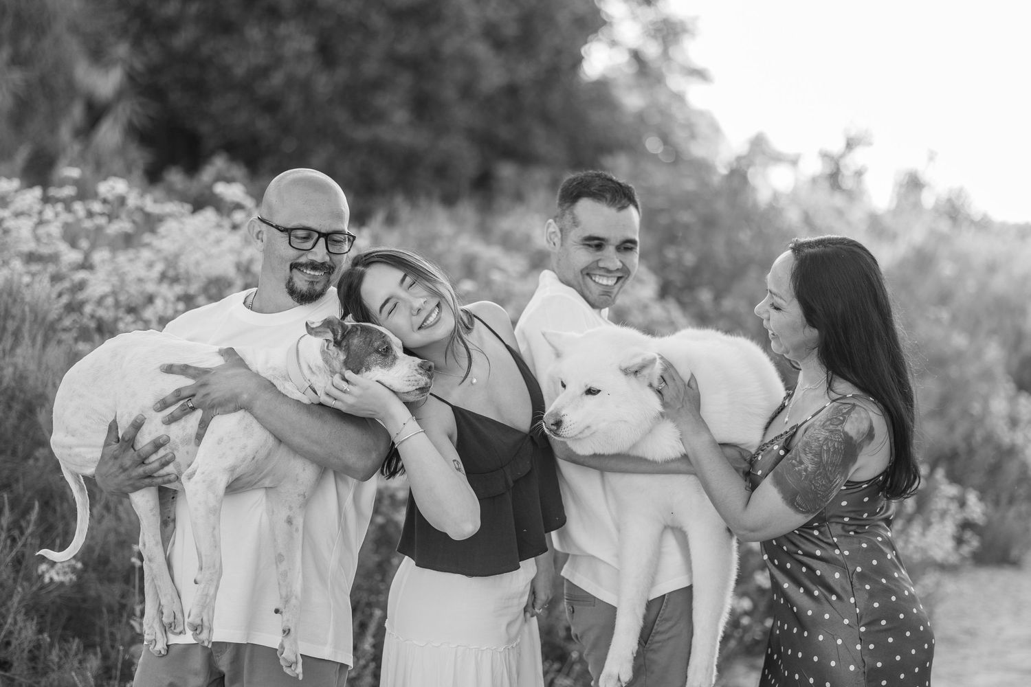 Black and white family photo of four adults joyfully holding white dogs outdoors in a natural setting.