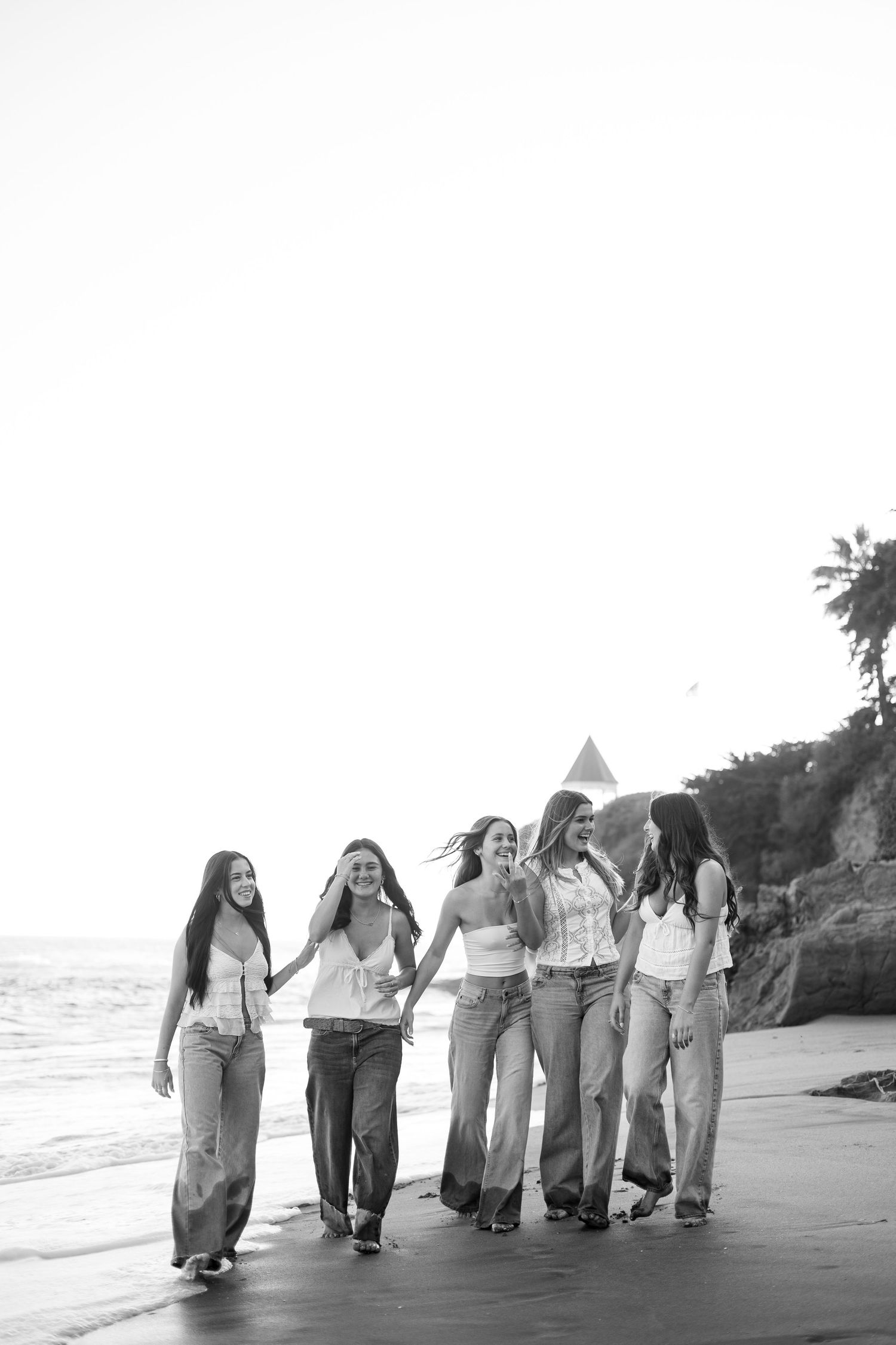 Black and white photo of six young women walking together on a beach, laughing and holding hands near the ocean.