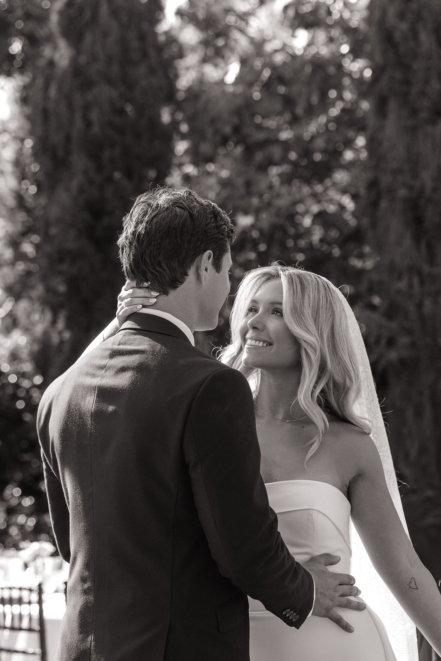 Black and white photo of a smiling blonde bride in a white dress embracing a groom in a suit outdoors.