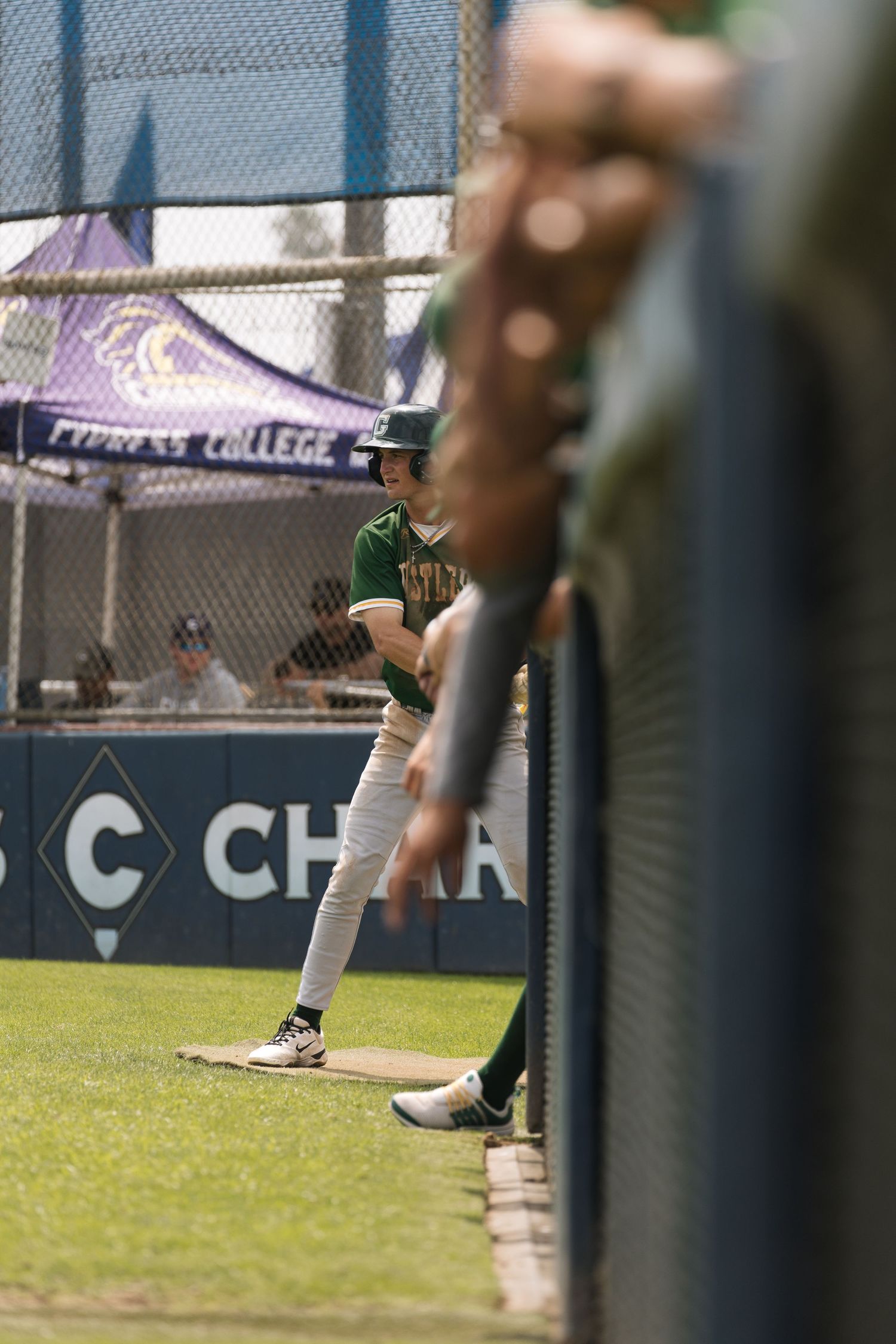 Baseball player in green uniform stands on base at Cypress College field during a sunny day game.