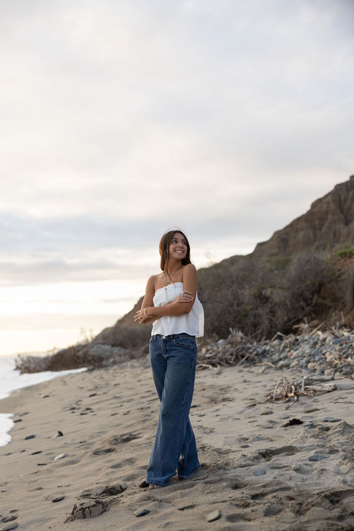 A smiling woman in a white top and flared jeans stands on a sandy beach with cliffs in the background.