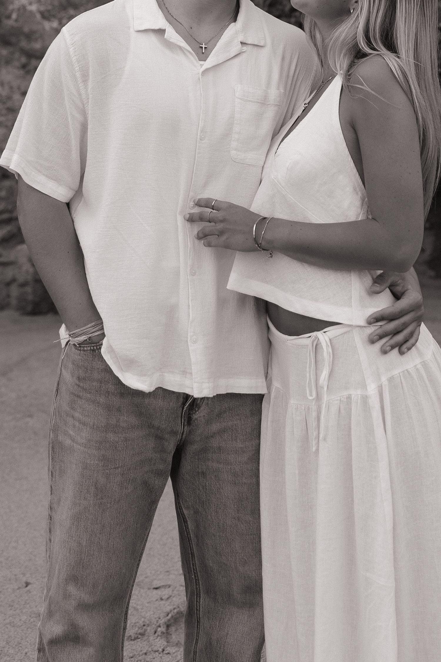 Black and white photo of a couple in white outfits embracing, man in linen shirt and jeans, woman in flowing dress.