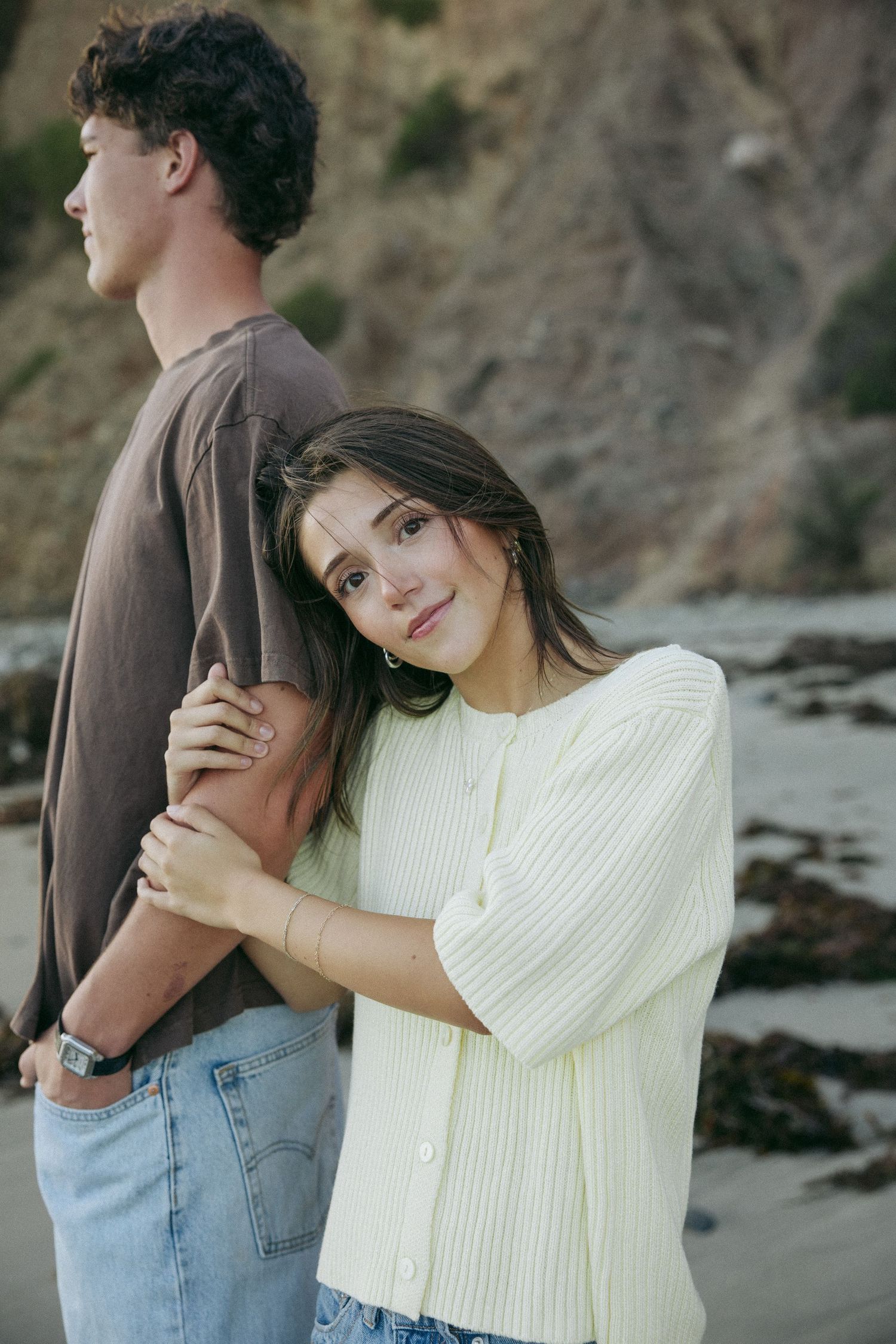 Young couple embracing on a rocky beach at sunset, the woman in a cream knit top leaning on the arm of a man in a brown t-shirt.