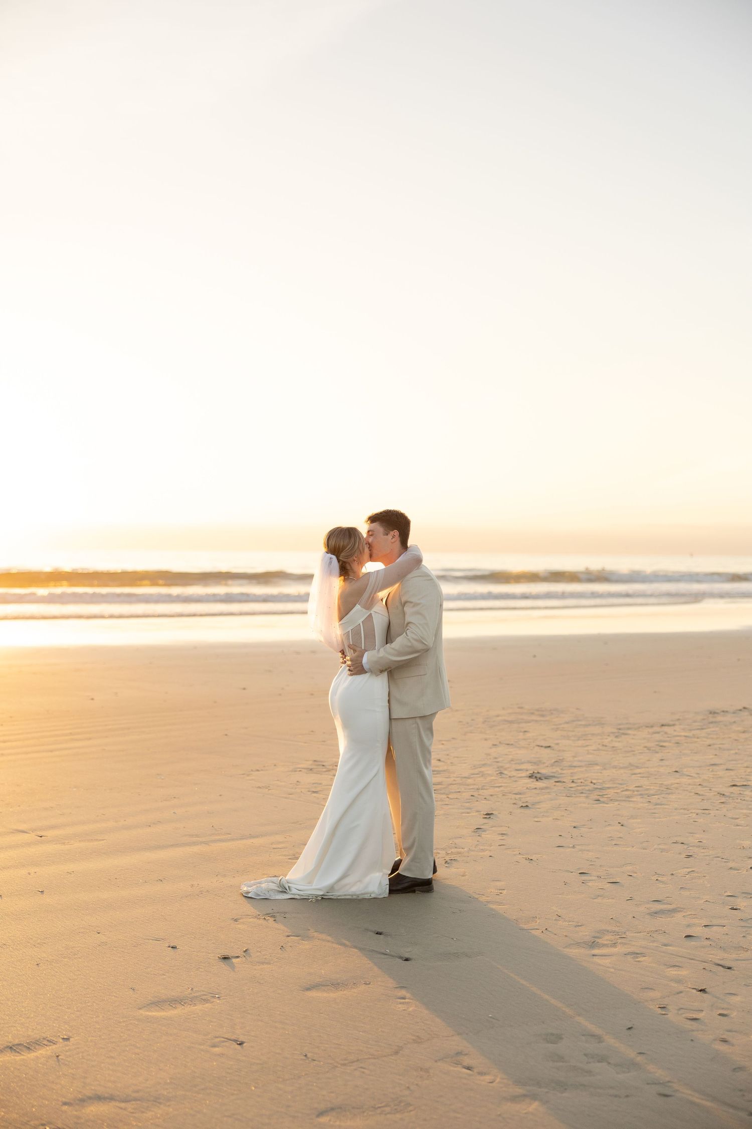 Bride and groom share a romantic kiss on a sandy beach at golden sunset, bride in white gown with veil.