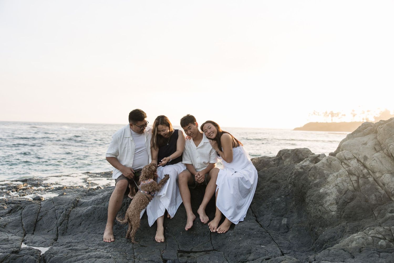 Family of four sitting on coastal rocks with a dog, dressed in white, enjoying a serene beach sunset together.