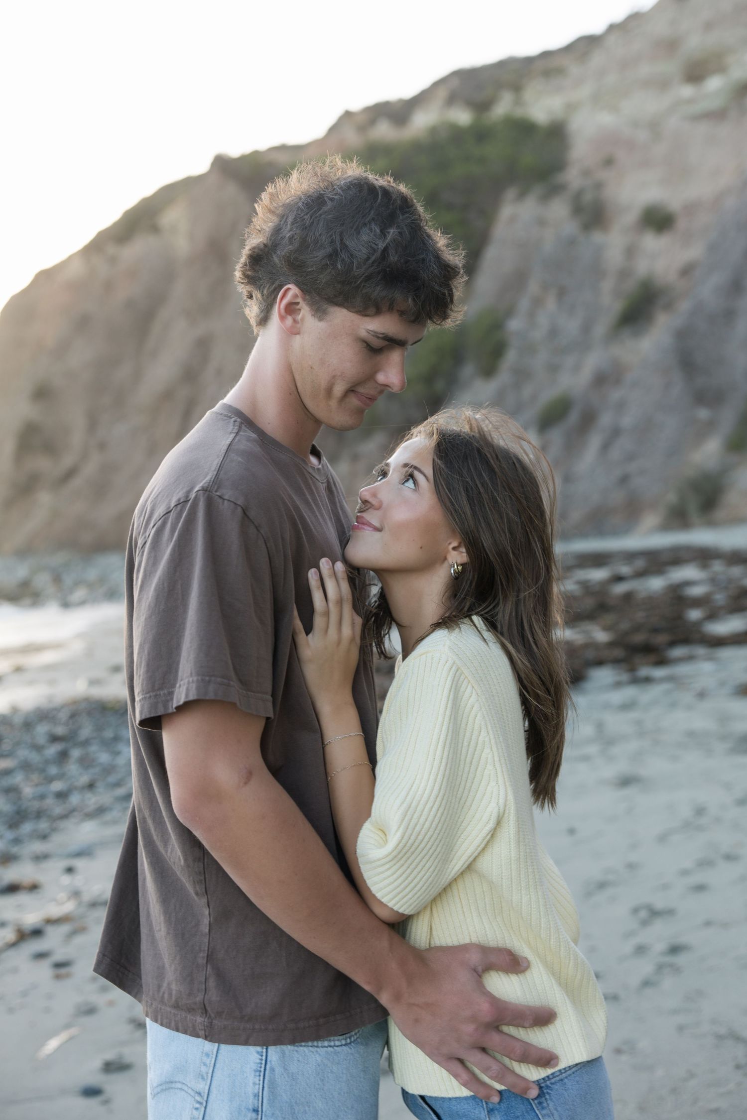 Young couple sharing a romantic moment on a rocky beach, embracing and gazing at each other at sunset.
