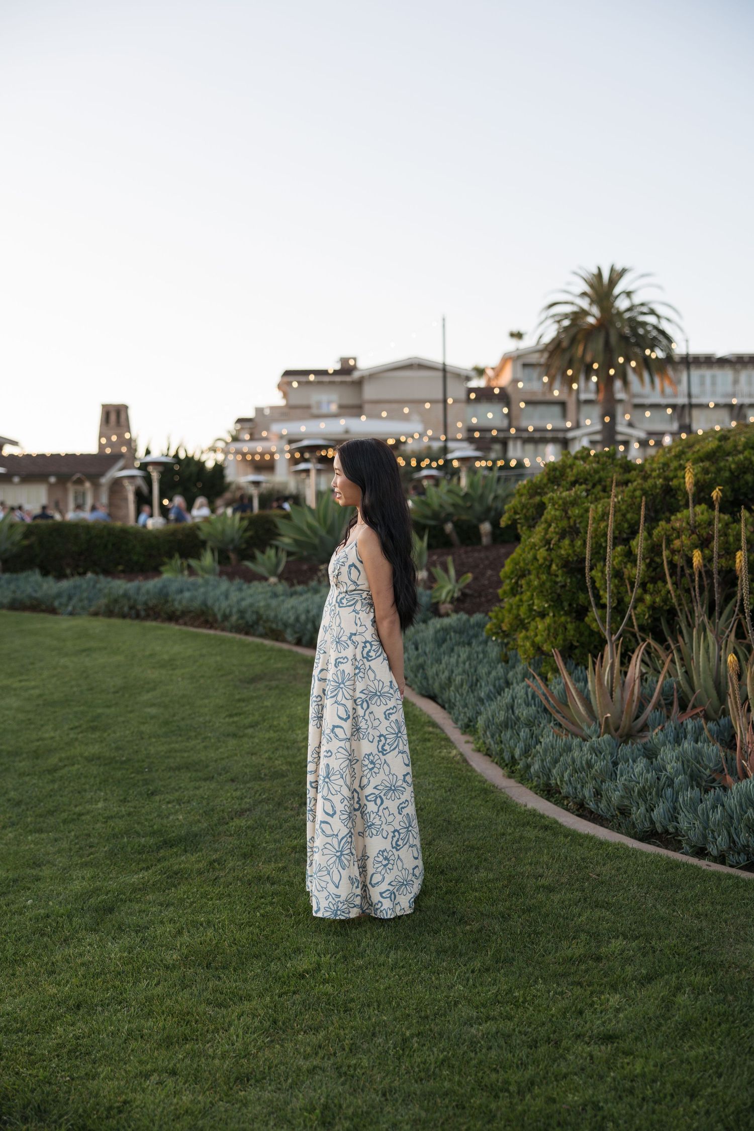 Woman in floral maxi dress standing on lush green lawn at dusk with garden and lit building backdrop.