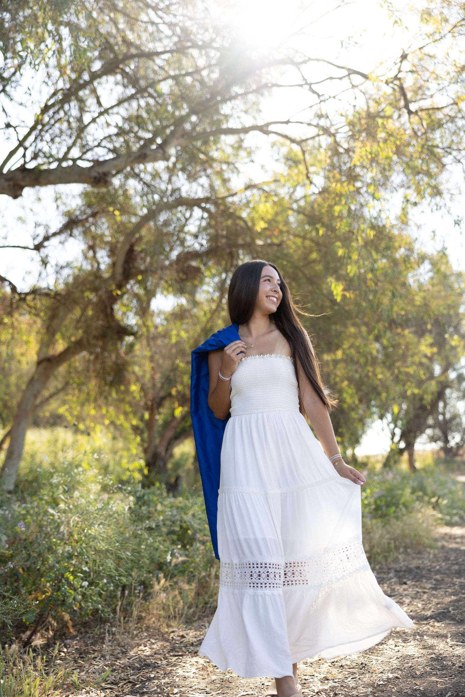 Smiling woman in white maxi dress holding blue jacket, standing in sunlit forest path.