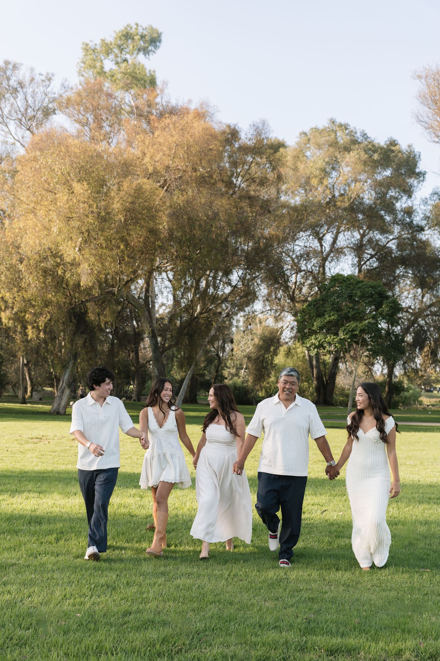 Family of five in white outfits holding hands and walking together across a green grass lawn with tall autumn trees in the background.
