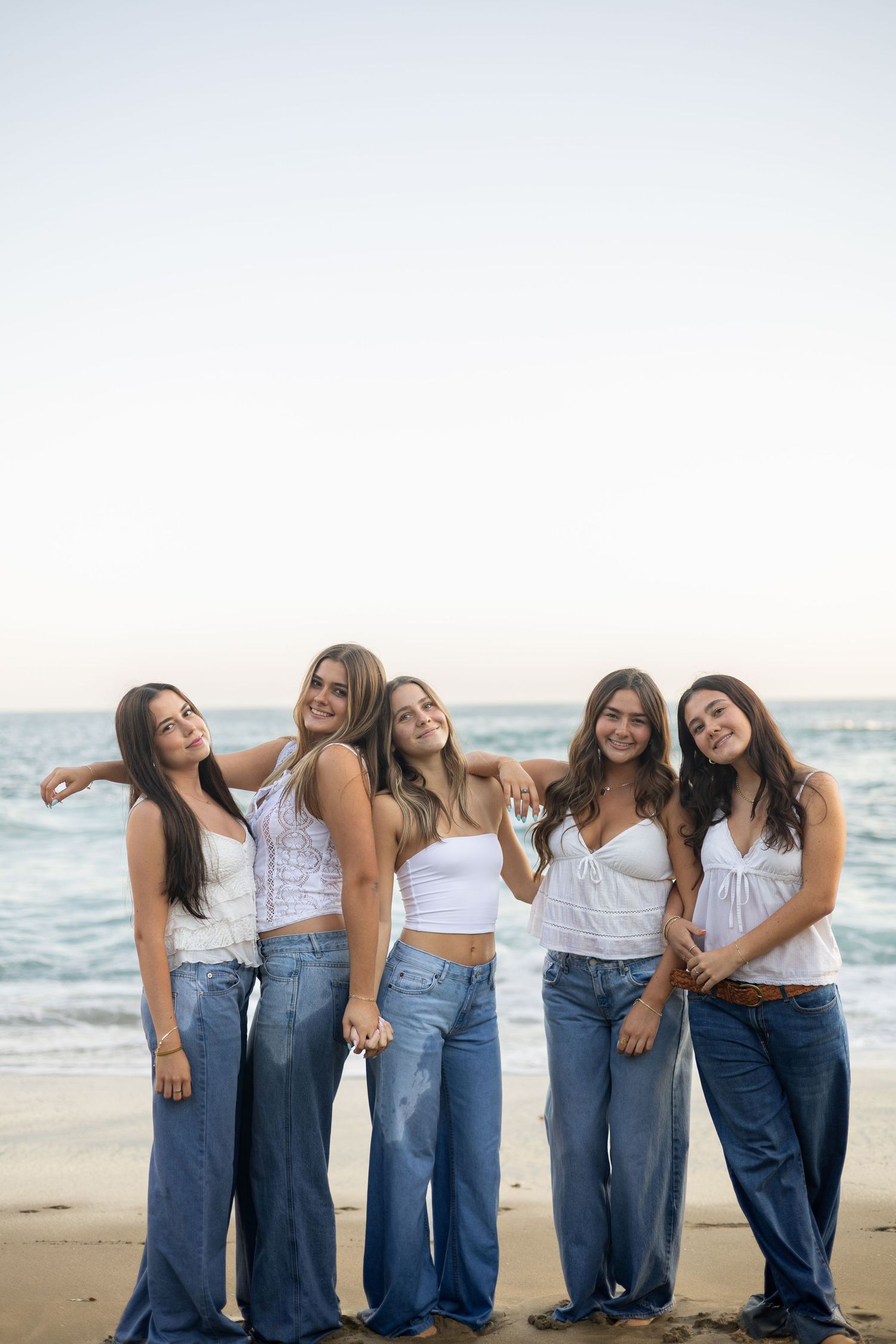 Friends in matching white tops and jeans pose playfully together on a beach.