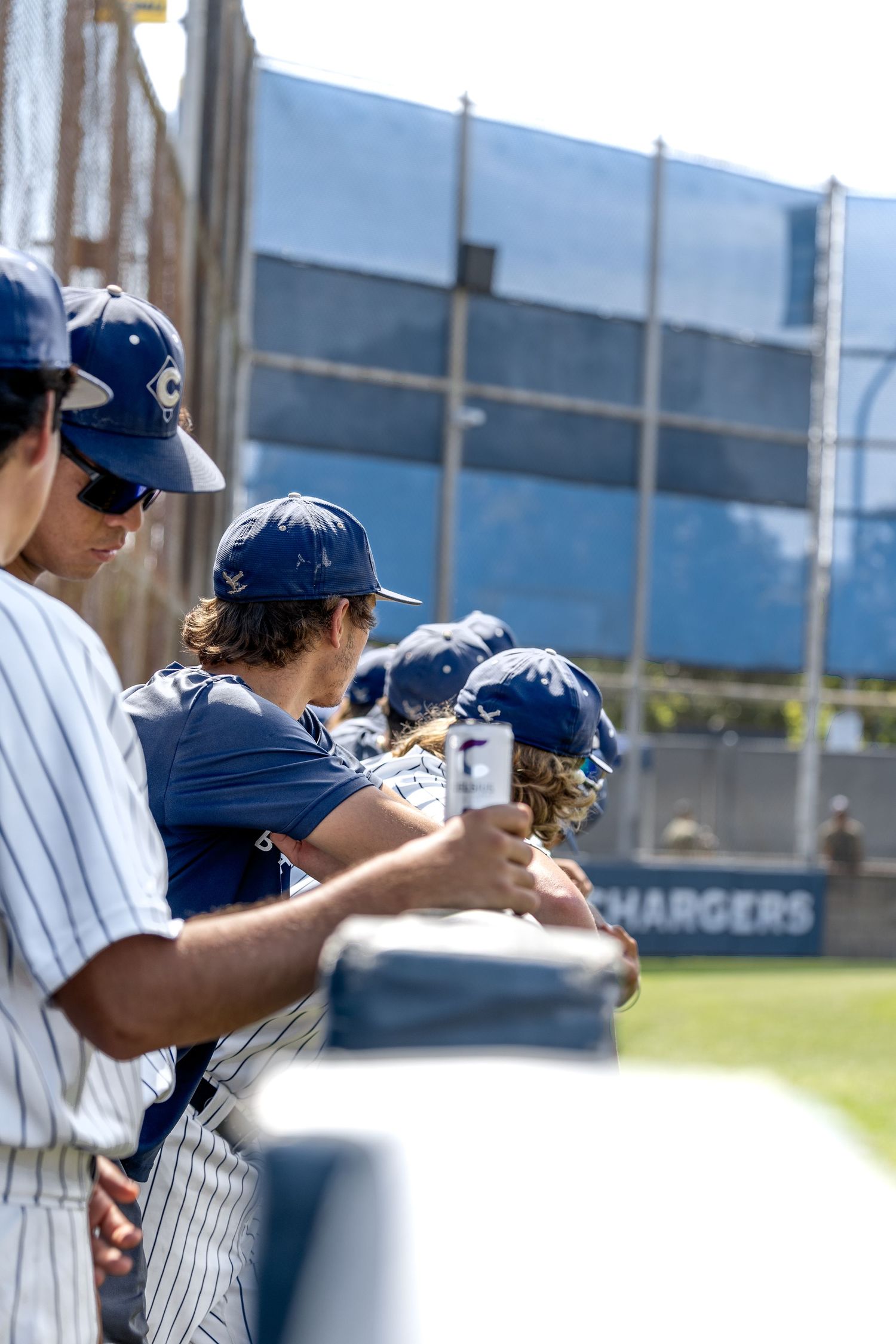 Baseball players in navy uniforms and caps stand in the dugout during a sunny game at Chargers baseball field.
