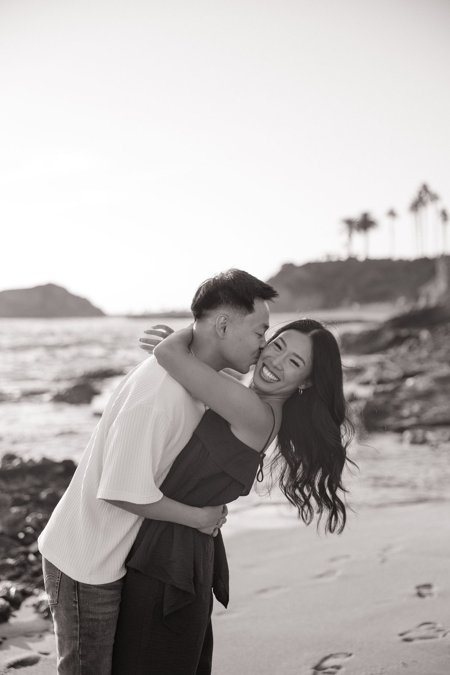 Black and white photo of a couple embracing and laughing on a rocky beach with palm trees and the ocean in the background.
