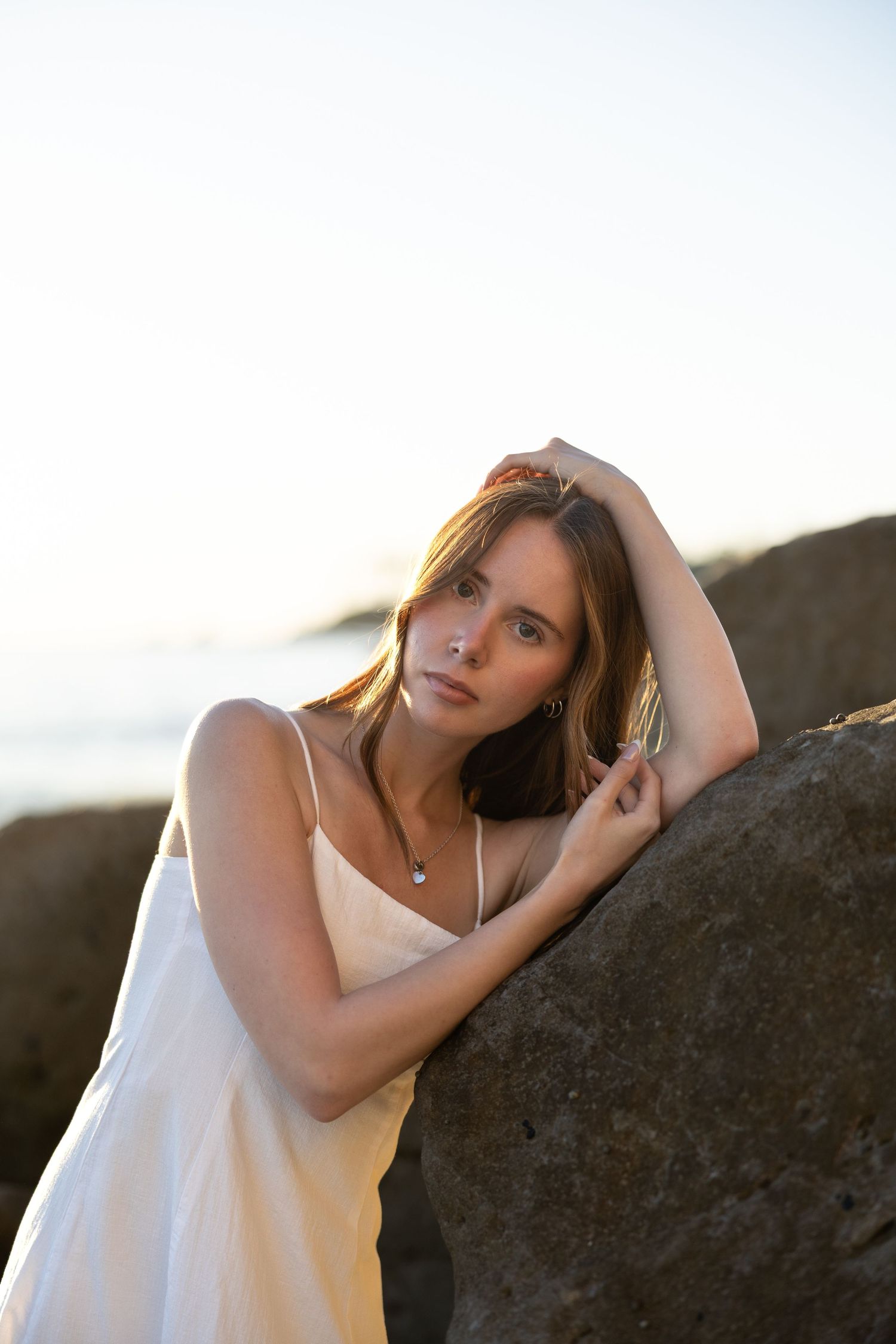 Young woman in white sundress leaning against coastal rocks at sunset, gazing at camera with serene expression.