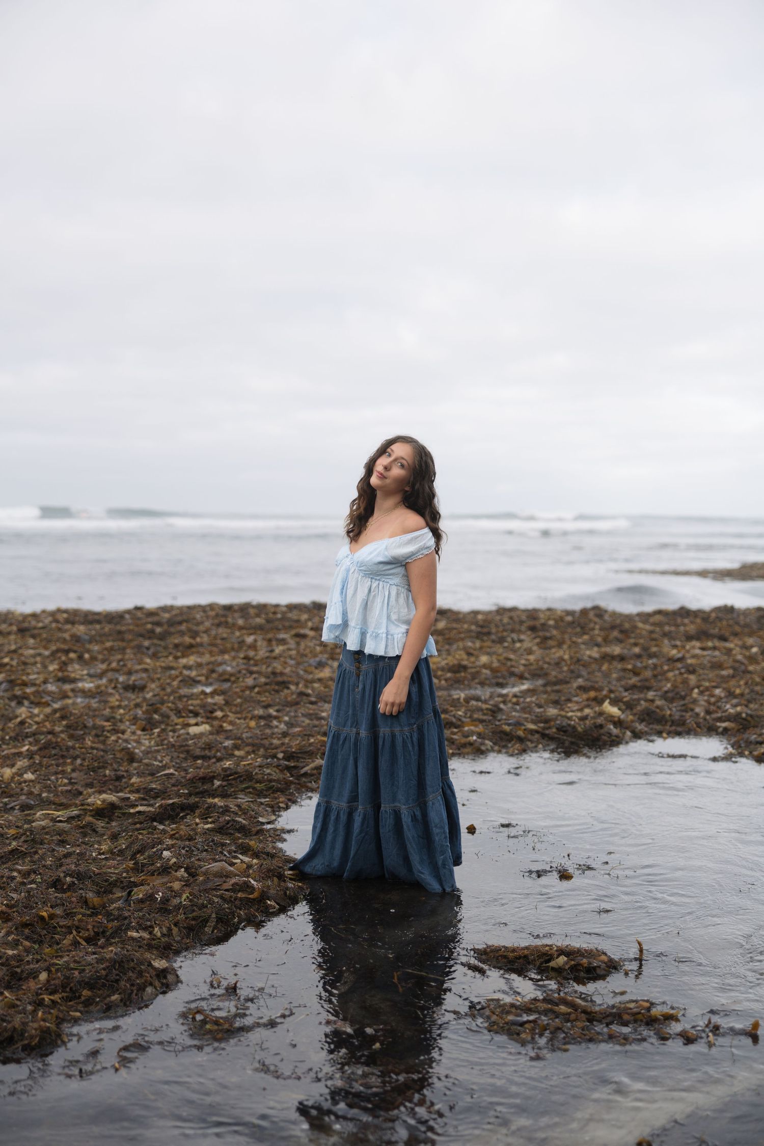 Woman in blue off-shoulder top and maxi skirt standing in shallow water on a rocky beach with ocean waves behind her.