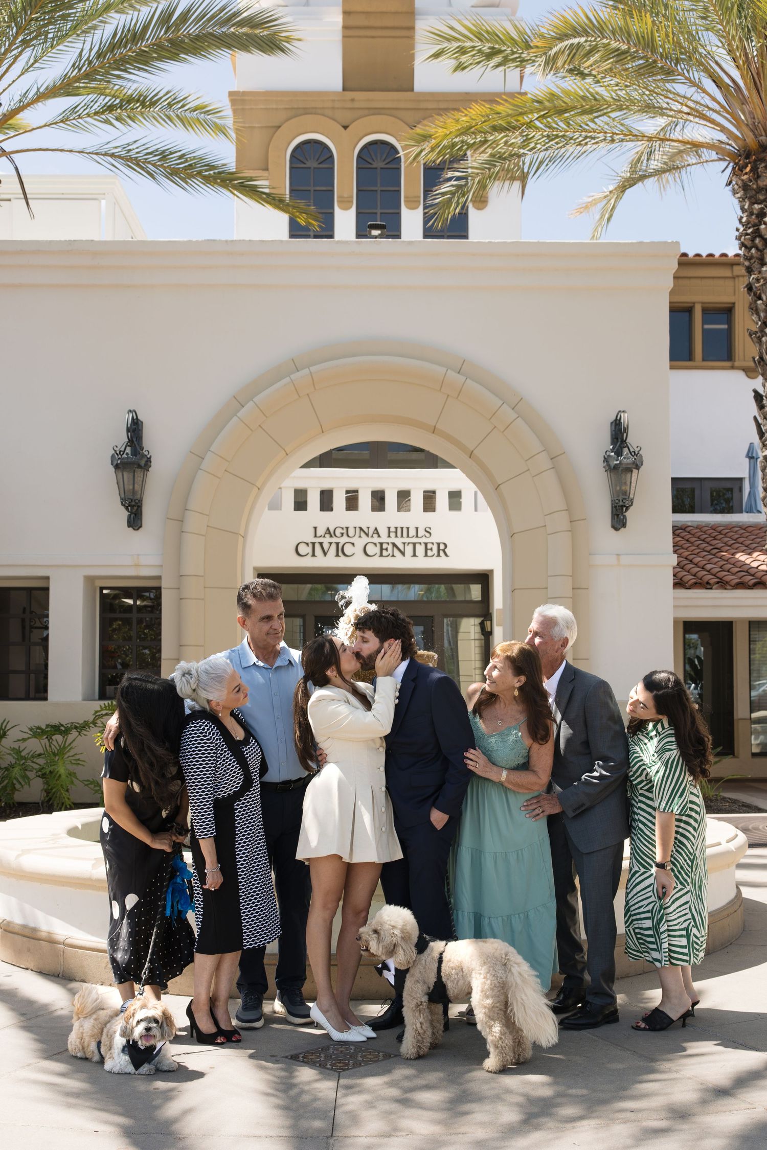 Newlywed couple kissing outside Laguna Hills Civic Center, surrounded by family and two fluffy dogs under palm trees.
