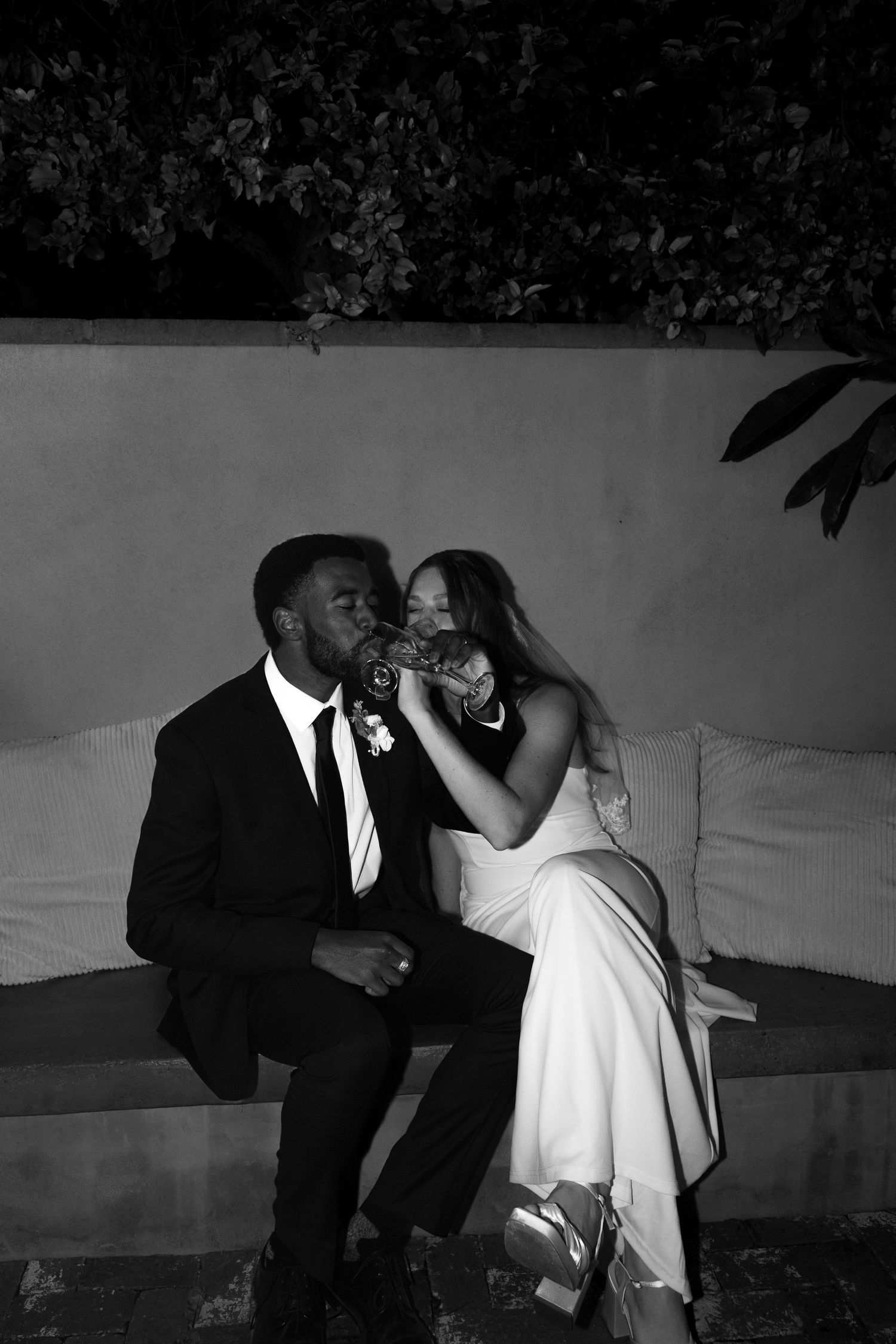 Black and white photo of a bride and groom sitting outdoors, sharing a drink together on their wedding night.