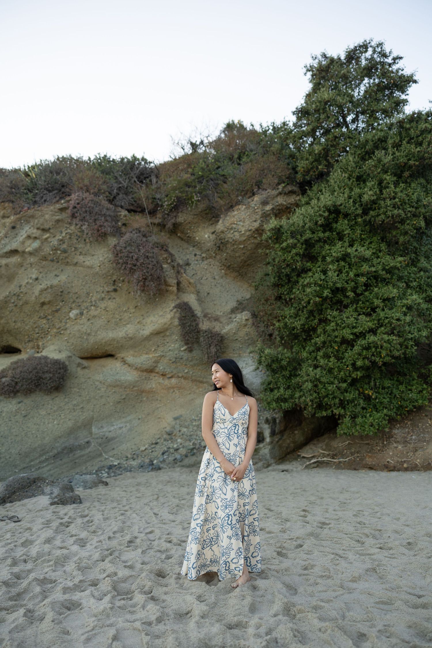 Woman in floral maxi dress standing on sandy beach with rocky cliffs and green trees in background.