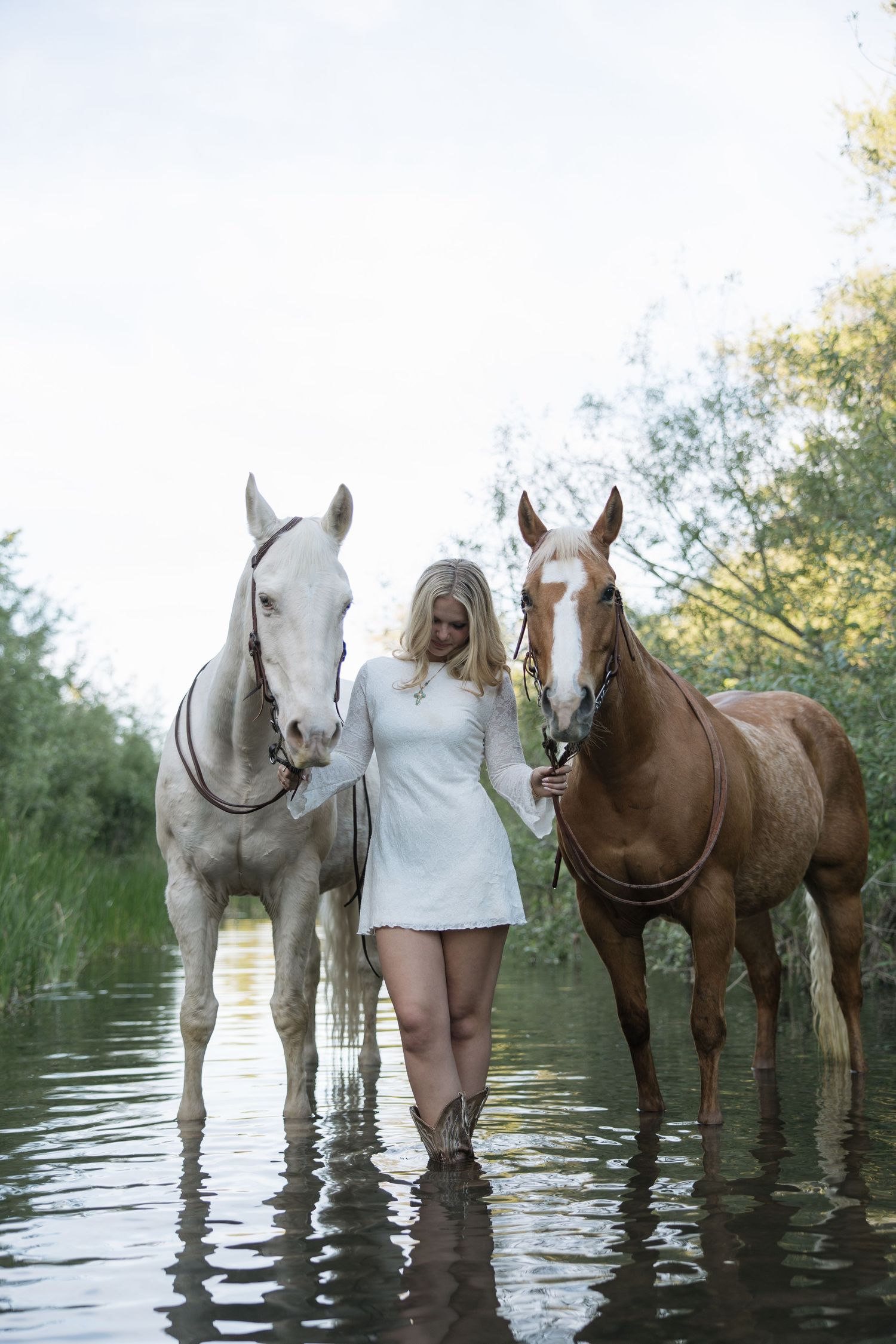 Woman in white dress standing in water with two horses, a white and a brown, surrounded by lush greenery.