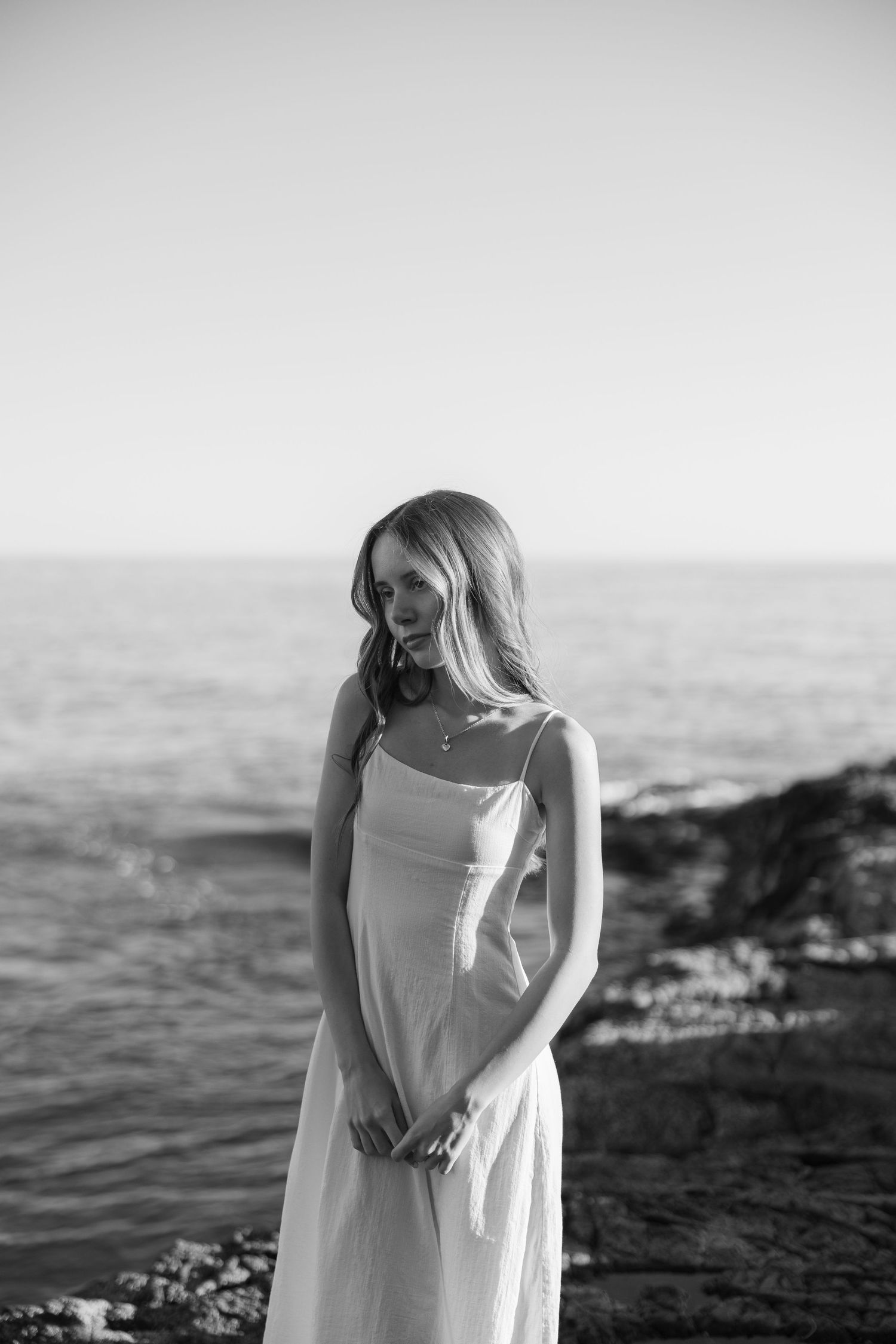 Black and white photo of a woman in a white sundress standing on rocky coastal shore with ocean in background.