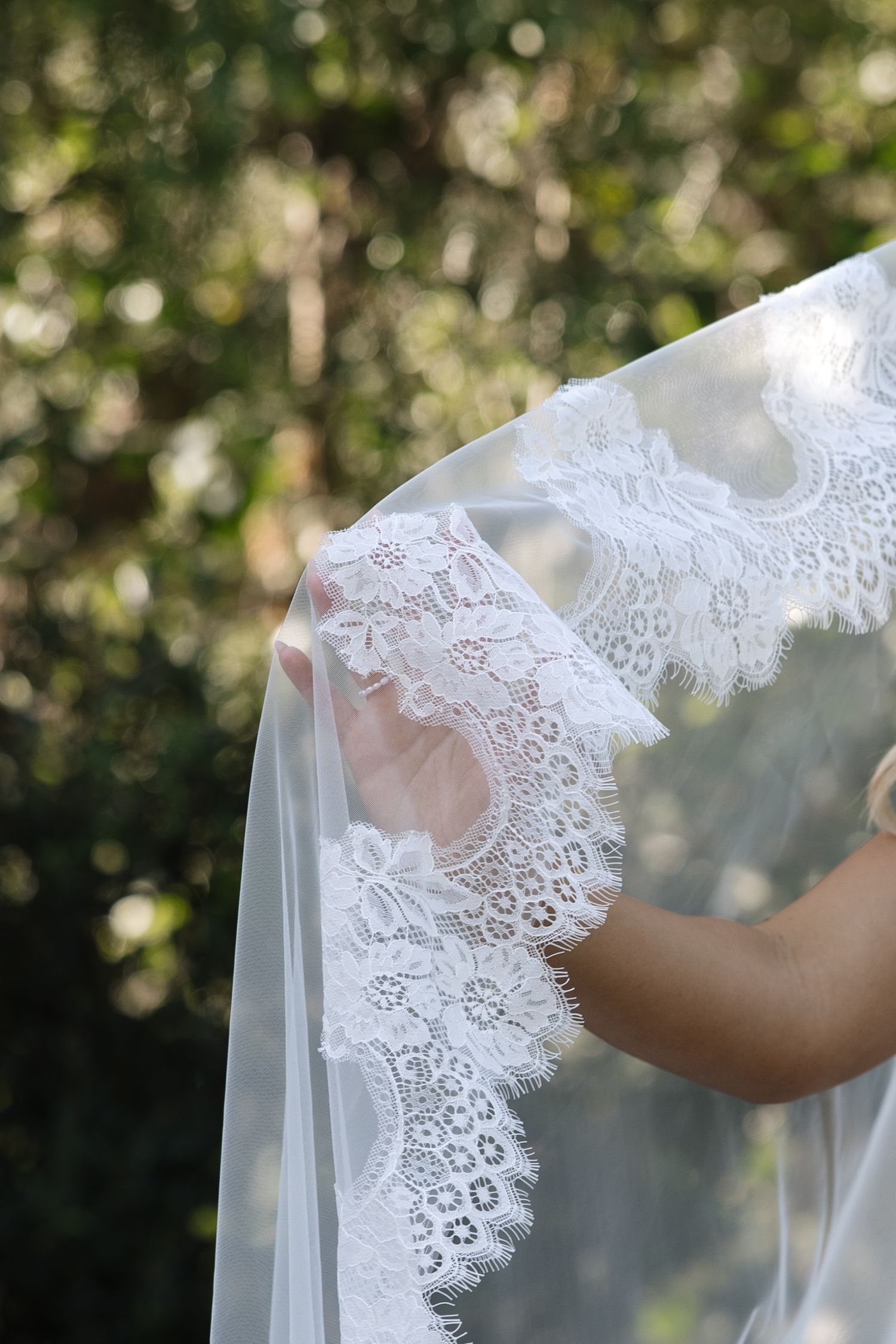 Bride holding a delicate white lace wedding veil outdoors with green foliage in the background.
