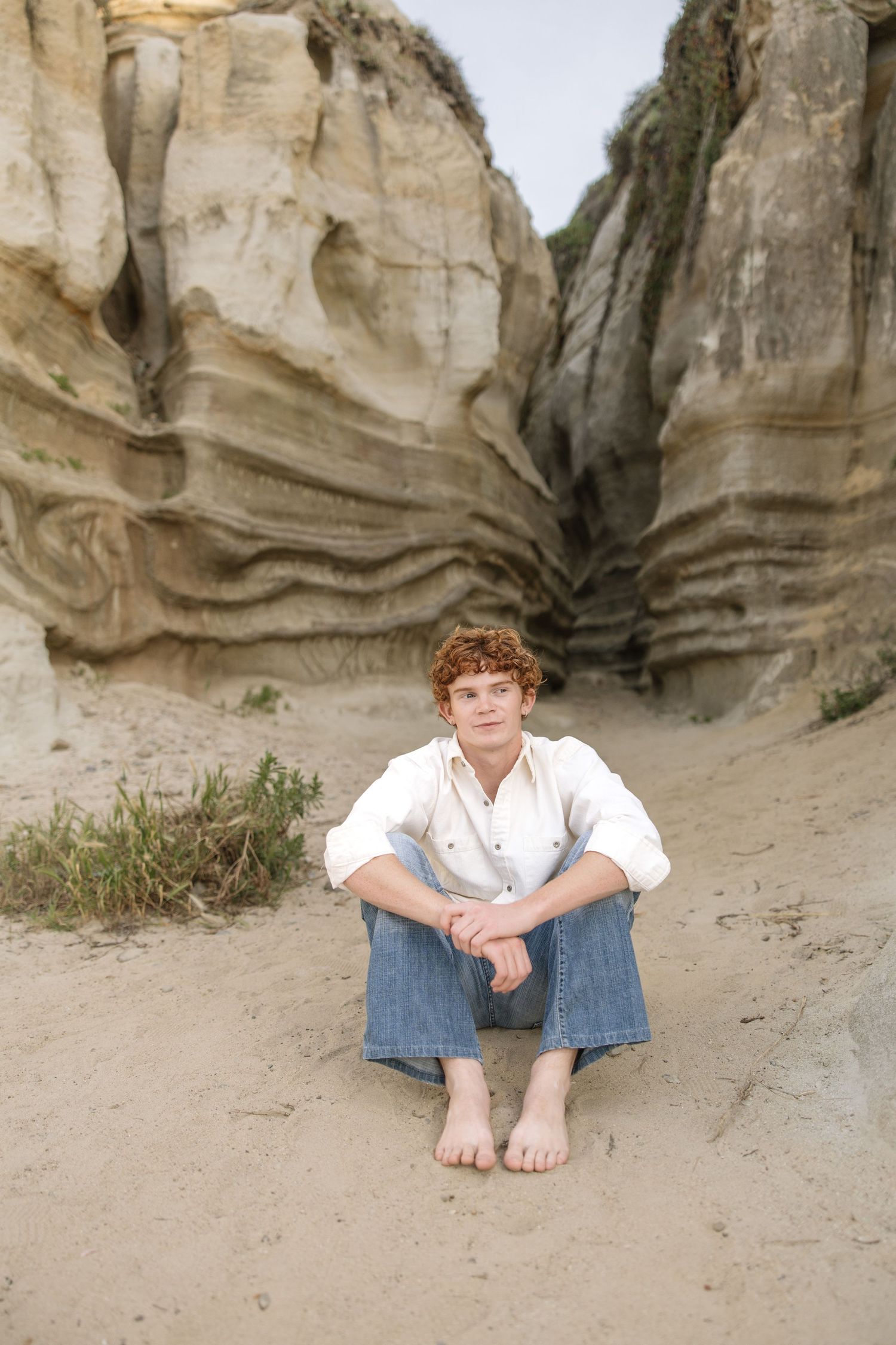 Young man with curly red hair sits barefoot on sandy ground between towering layered rock formations.
