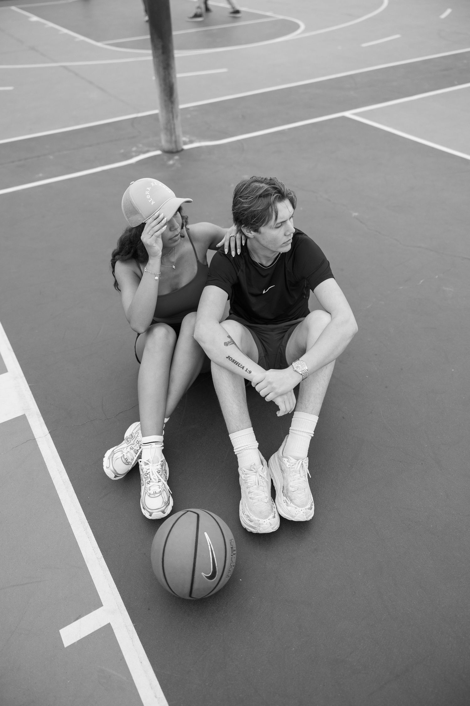 Two people sit casually on a basketball court with a ball nearby in black and white photography.