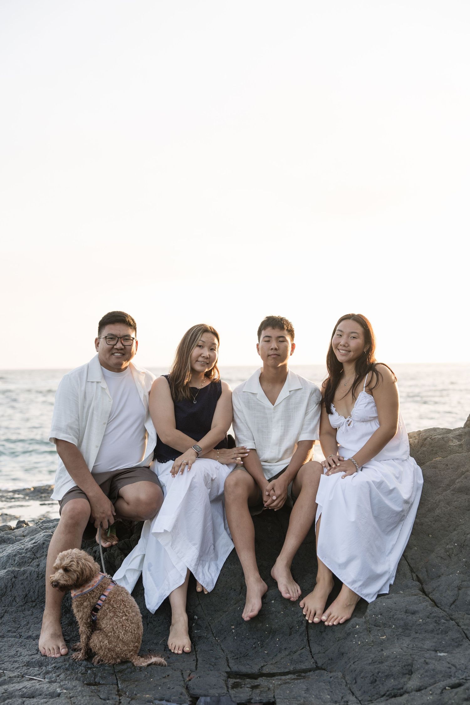 Four friends and a fluffy dog sit on coastal rocks at sunset, smiling at the camera near the ocean.