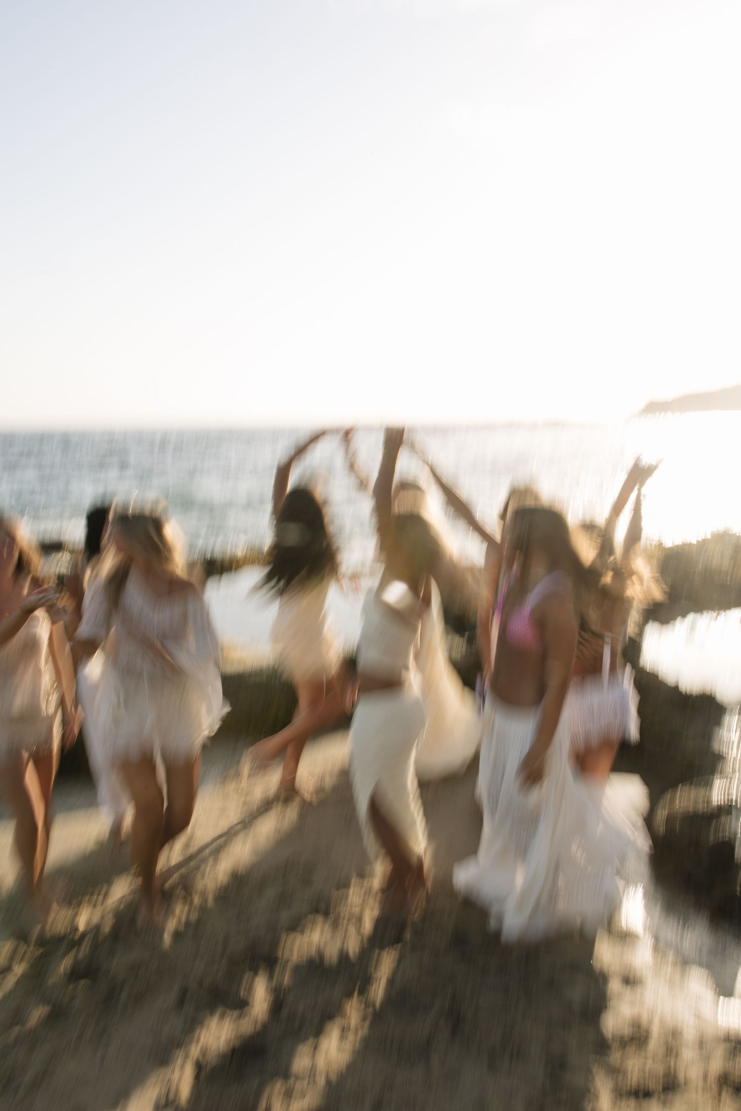 Blurred motion photo of women in white dresses dancing joyfully on a sunny beach by the ocean.