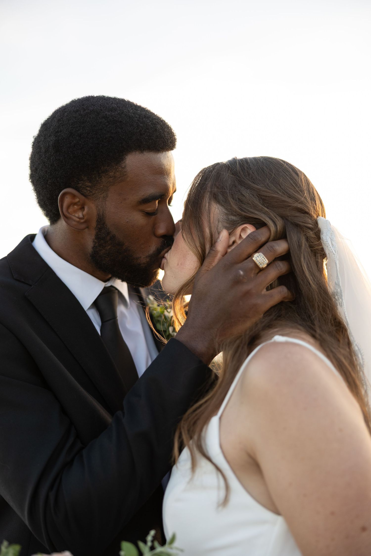 A groom in a black suit tenderly kisses his bride in a white dress and veil during their wedding ceremony.