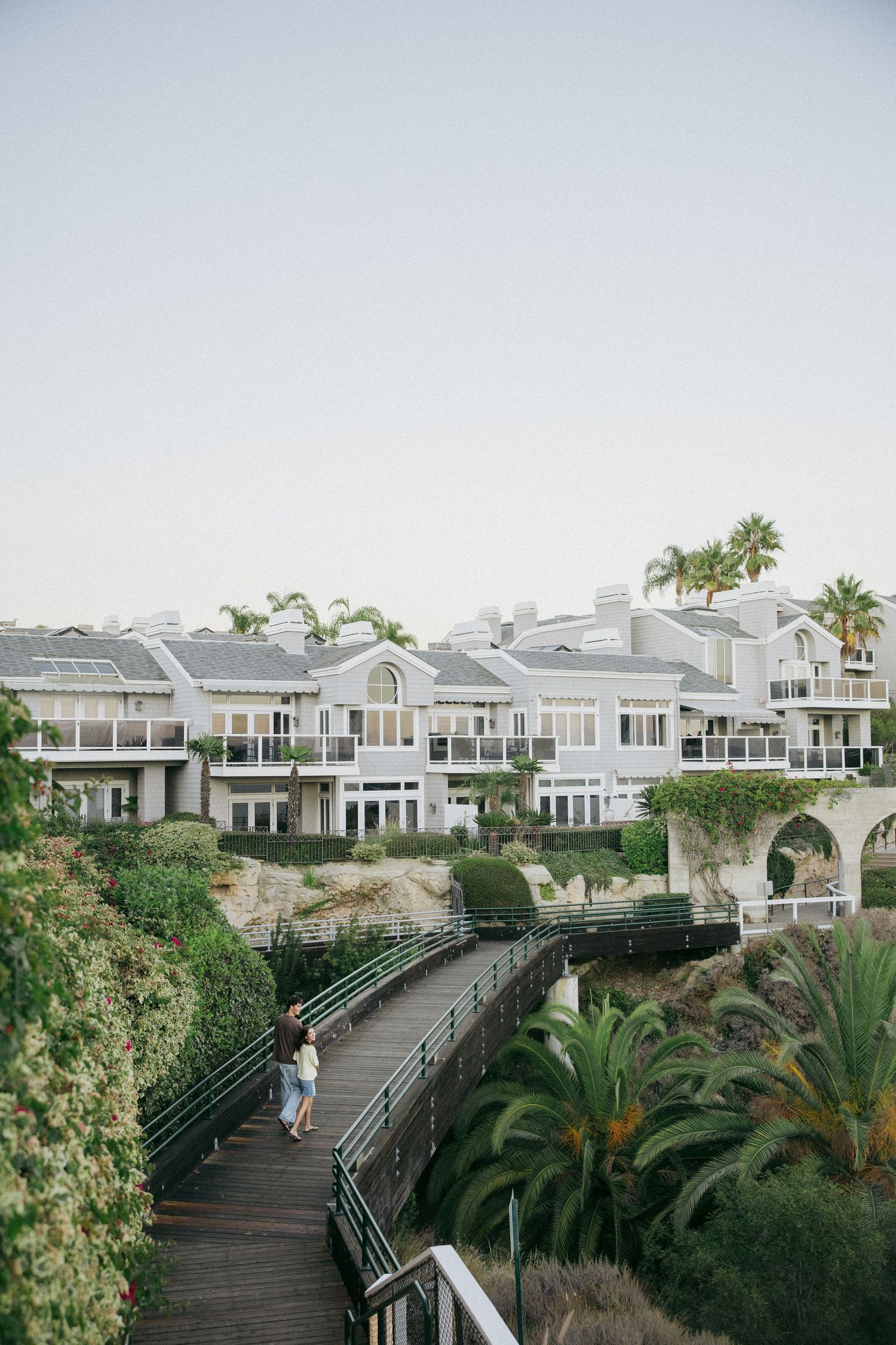 Couple walks along a curved wooden boardwalk toward elegant white coastal villas surrounded by palm trees and lush greenery.