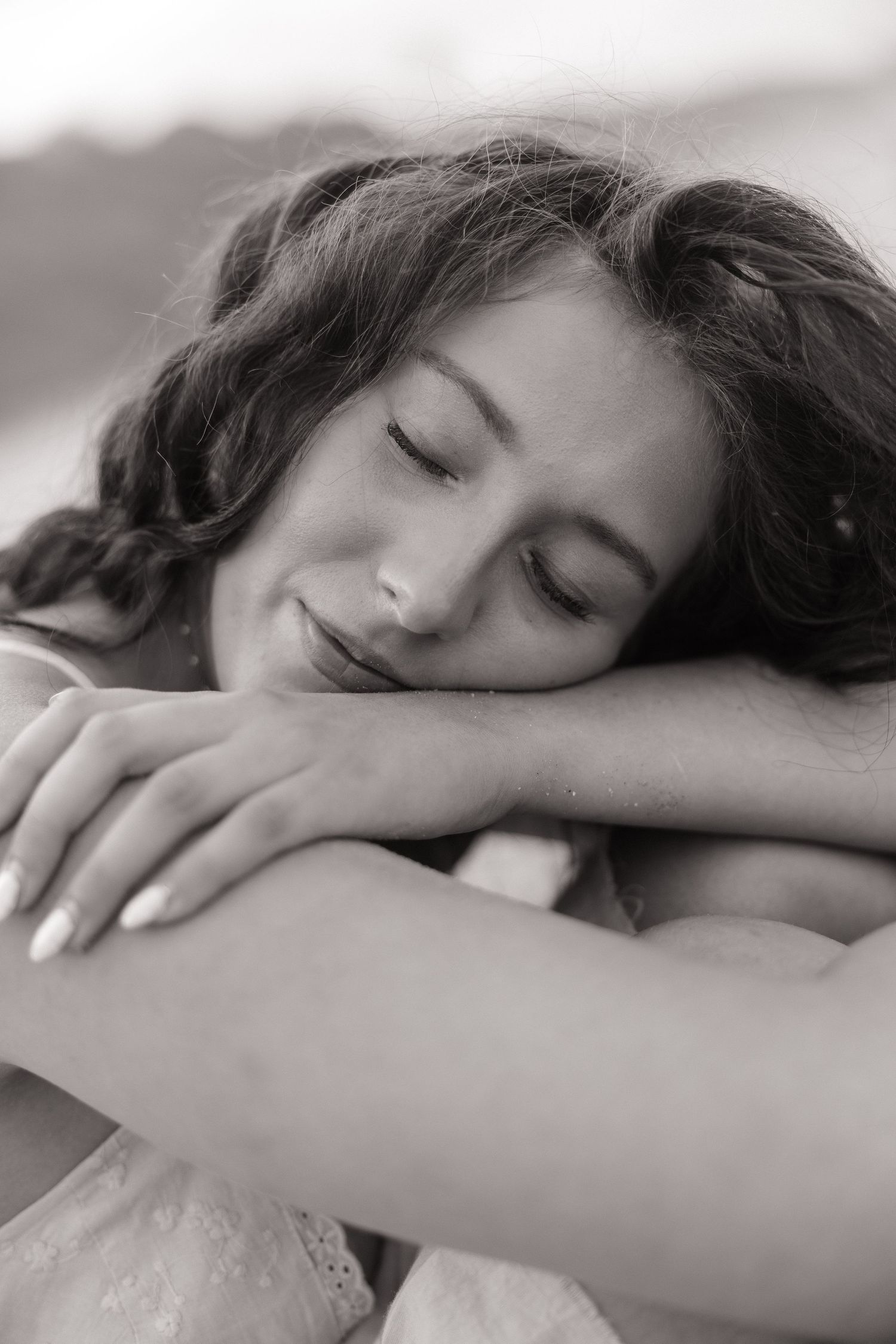 Black and white portrait of a young woman resting her head on her arms, eyes closed, smiling peacefully outdoors.