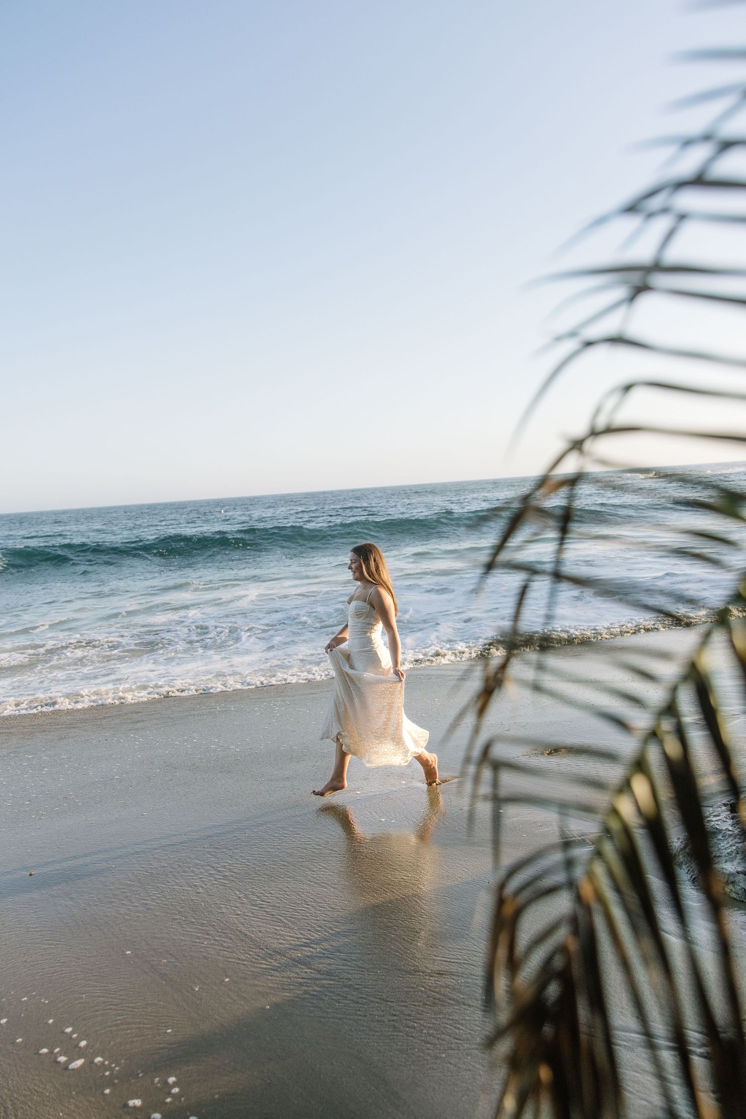 Woman in white dress walking barefoot on a sunny beach with ocean waves and palm fronds in foreground.