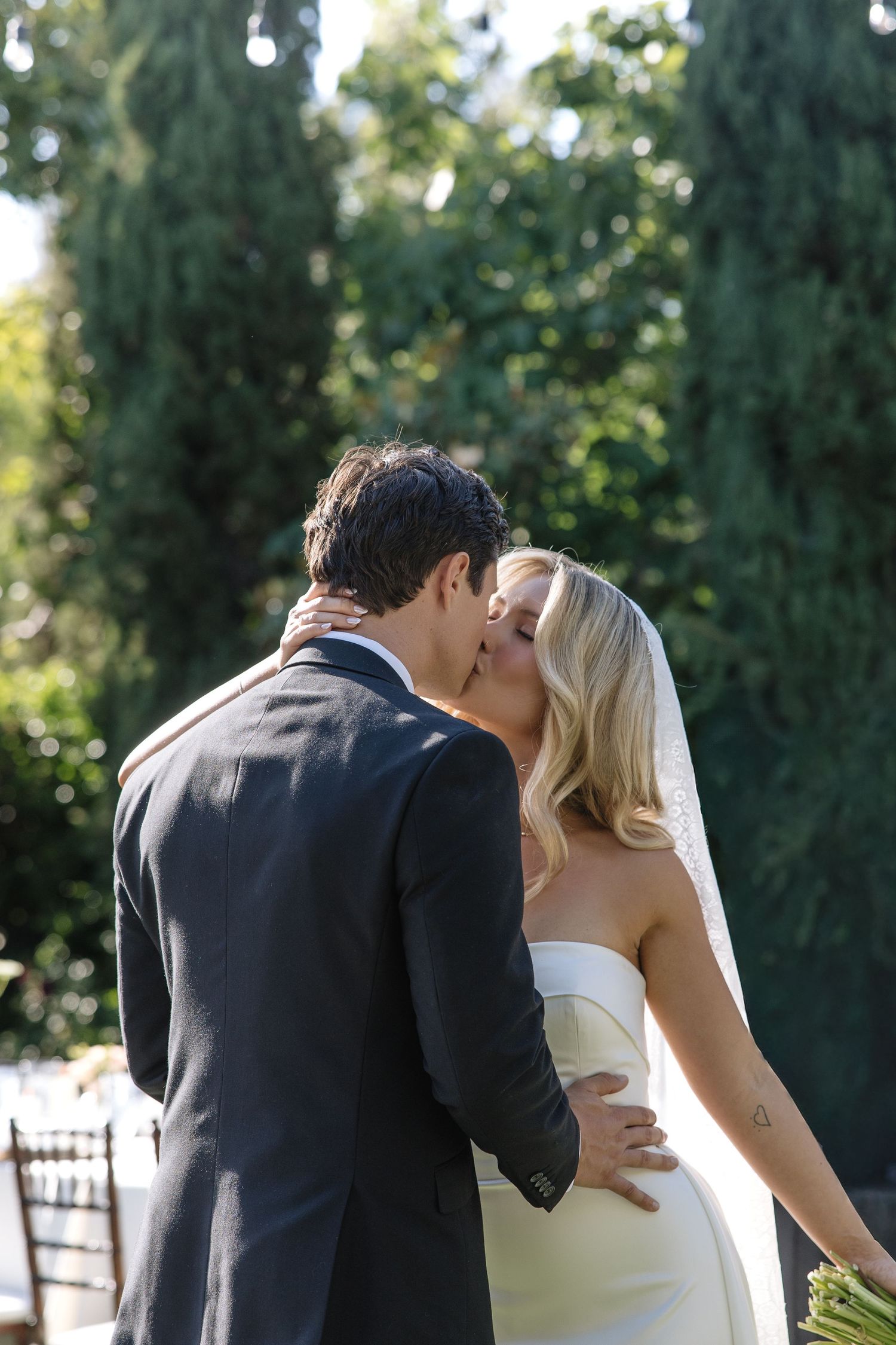 A bride and groom share a romantic kiss outdoors, surrounded by lush green trees on their wedding day.