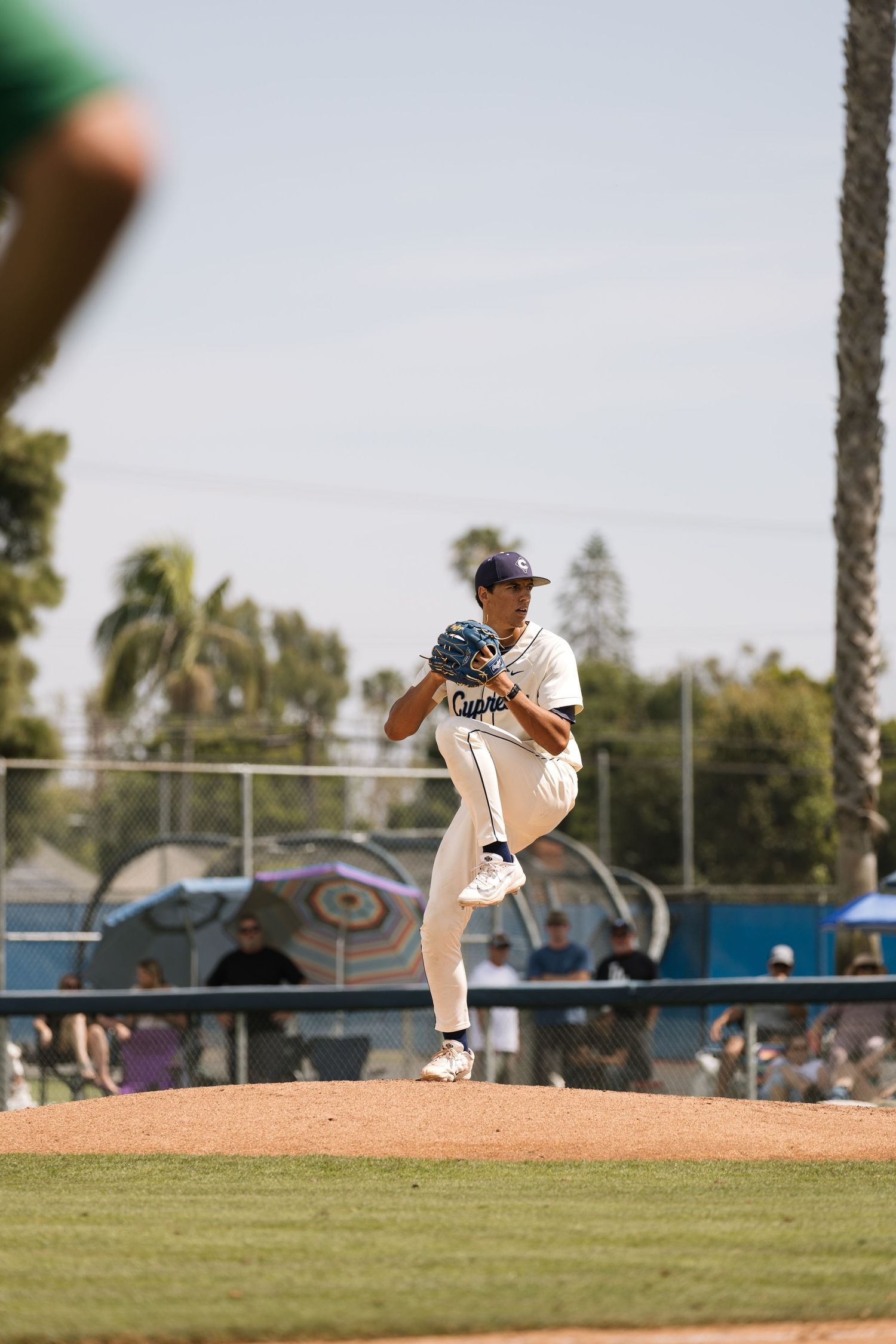 Baseball pitcher in white uniform mid-windup on mound during sunny outdoor game with palm trees in background.