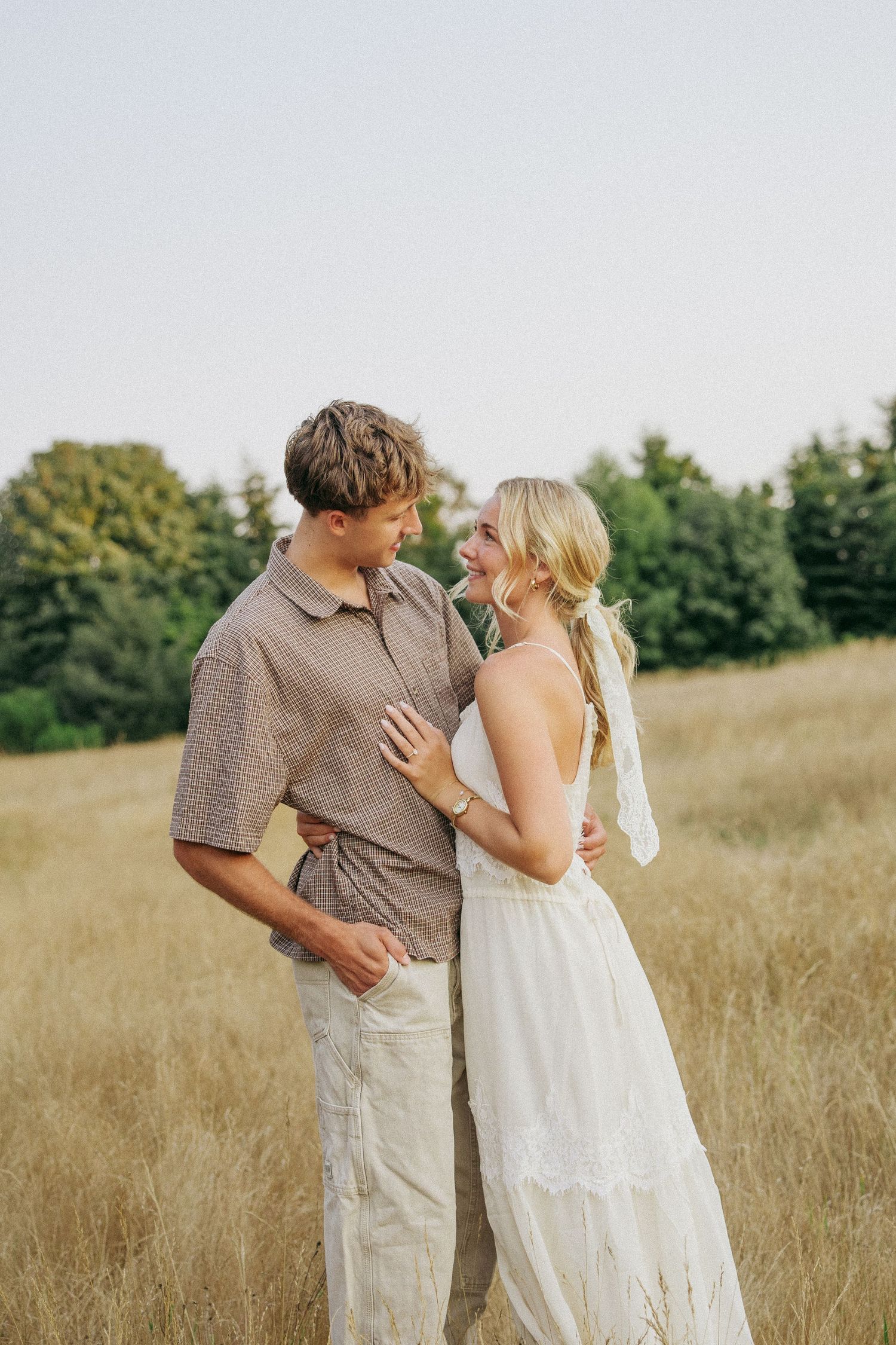 A couple shares a romantic moment in a golden field, the woman in a white dress and man in a brown shirt gazing at each other.