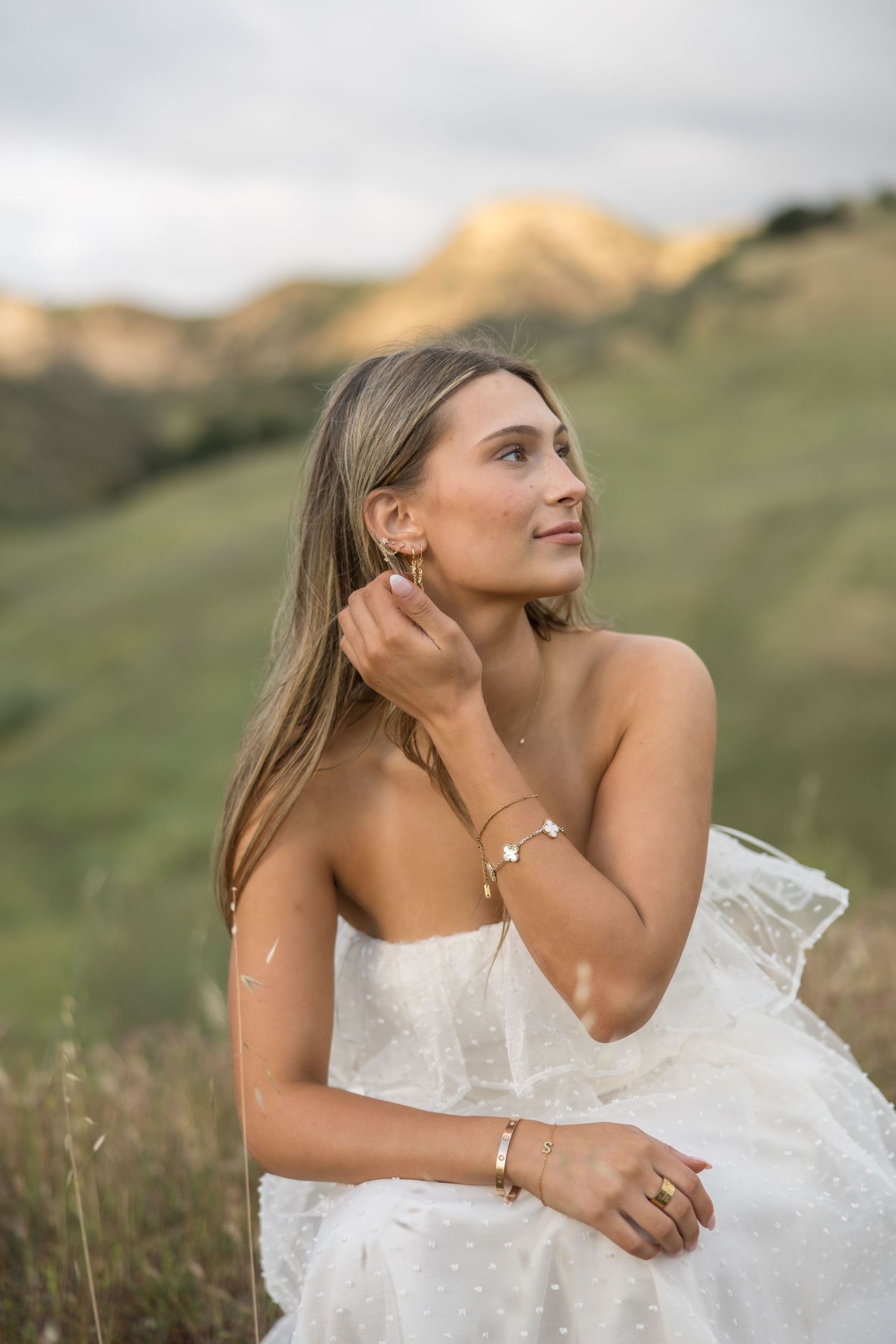 Woman in white strapless dress wearing gold jewelry, sitting in green hills at golden hour.