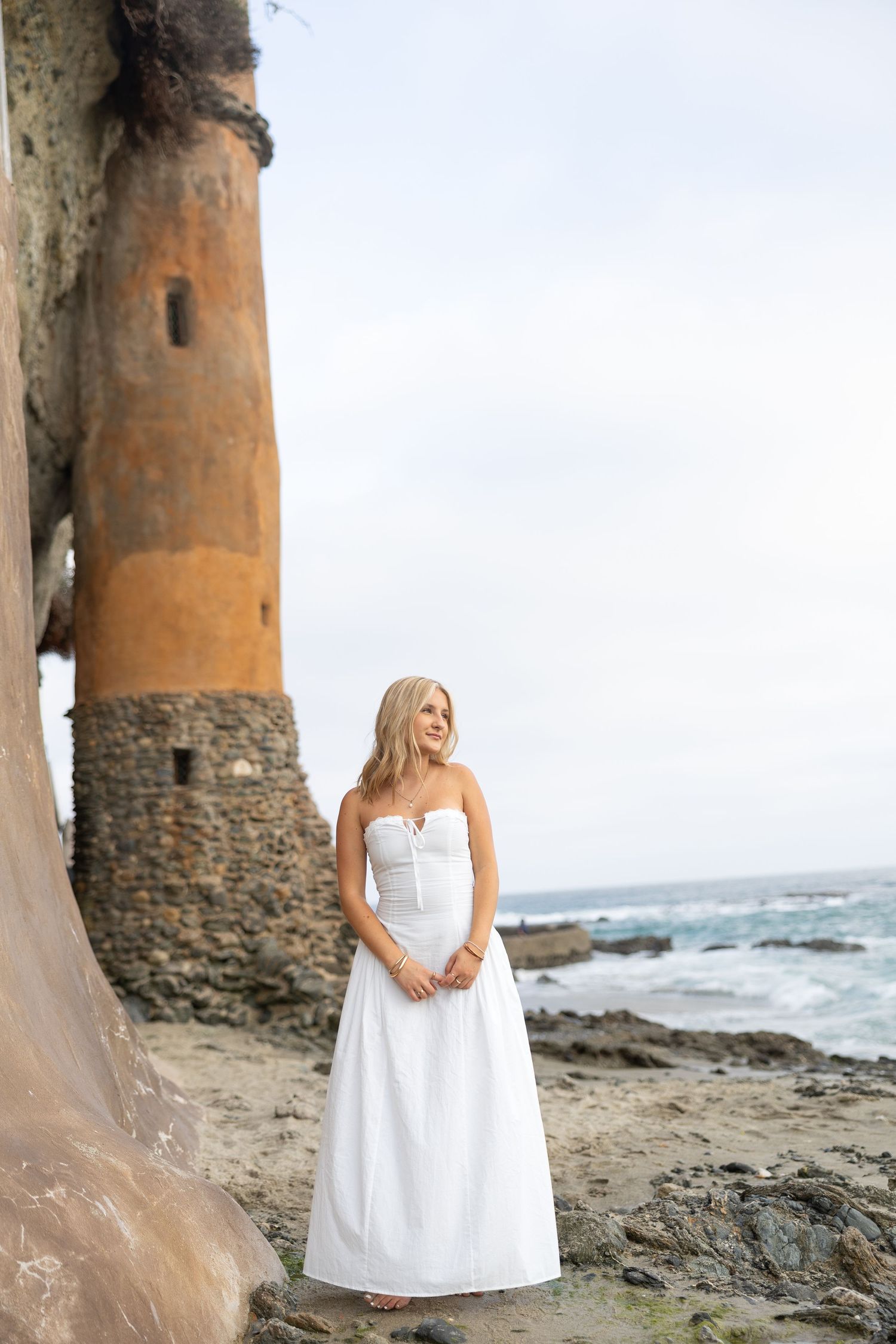 Blonde woman in white strapless gown standing on rocky beach near ancient stone tower by the ocean.