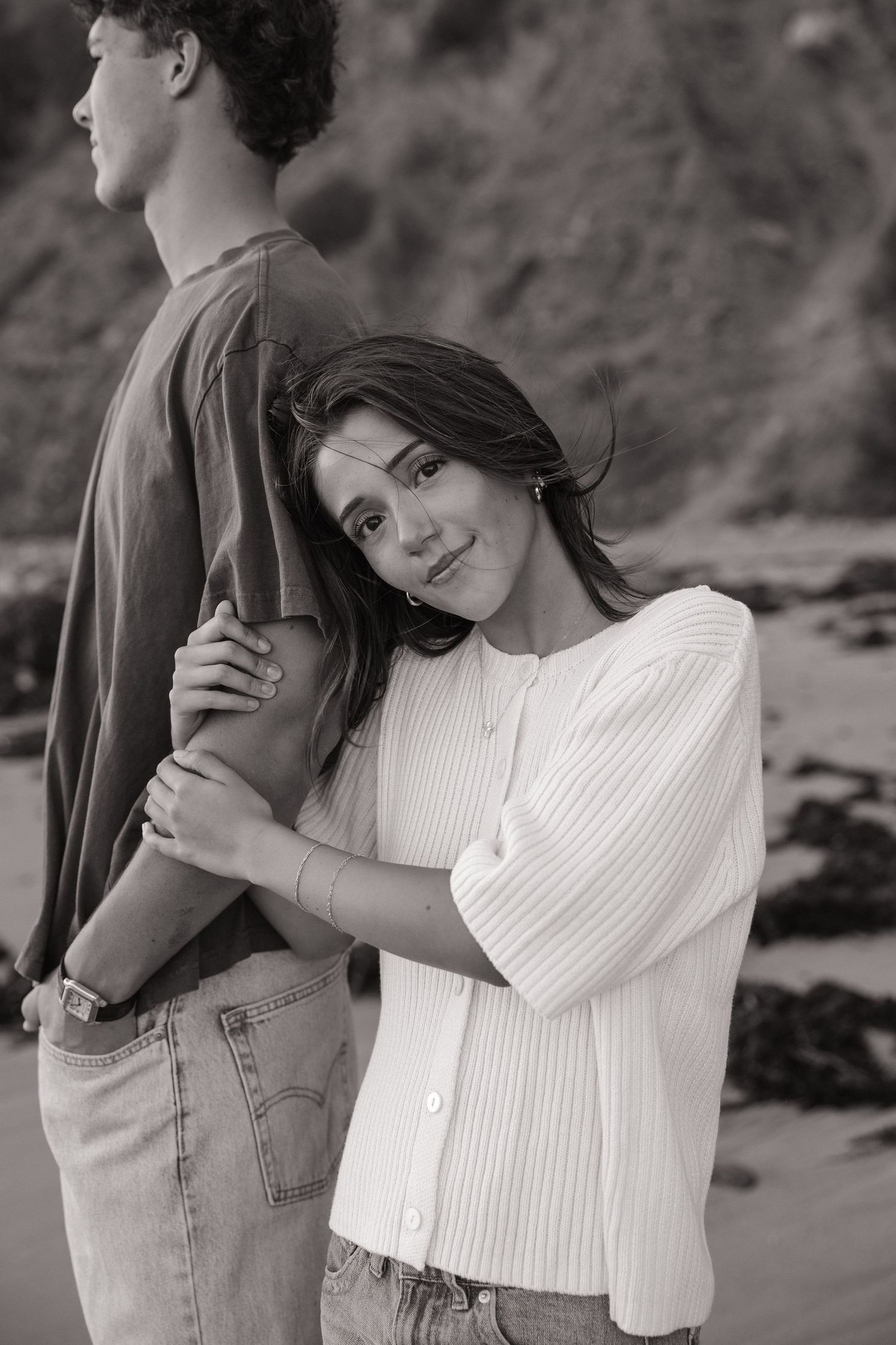 Black and white portrait of a young woman leaning her head on a tall man's arm while standing together on a rocky beach.