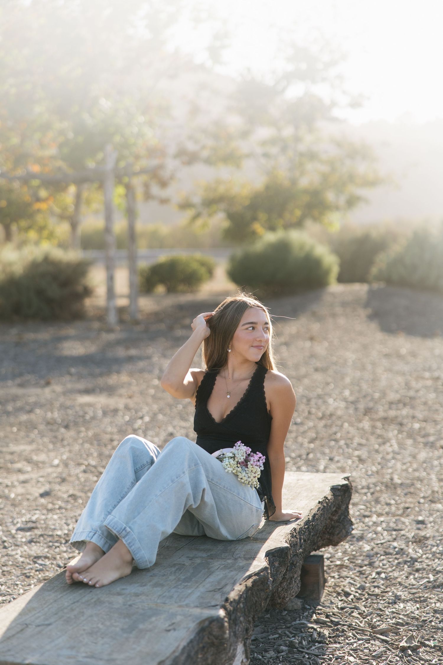 Young woman in black tank top and jeans sits barefoot on stone ledge holding wildflowers in soft golden sunlight outdoors.