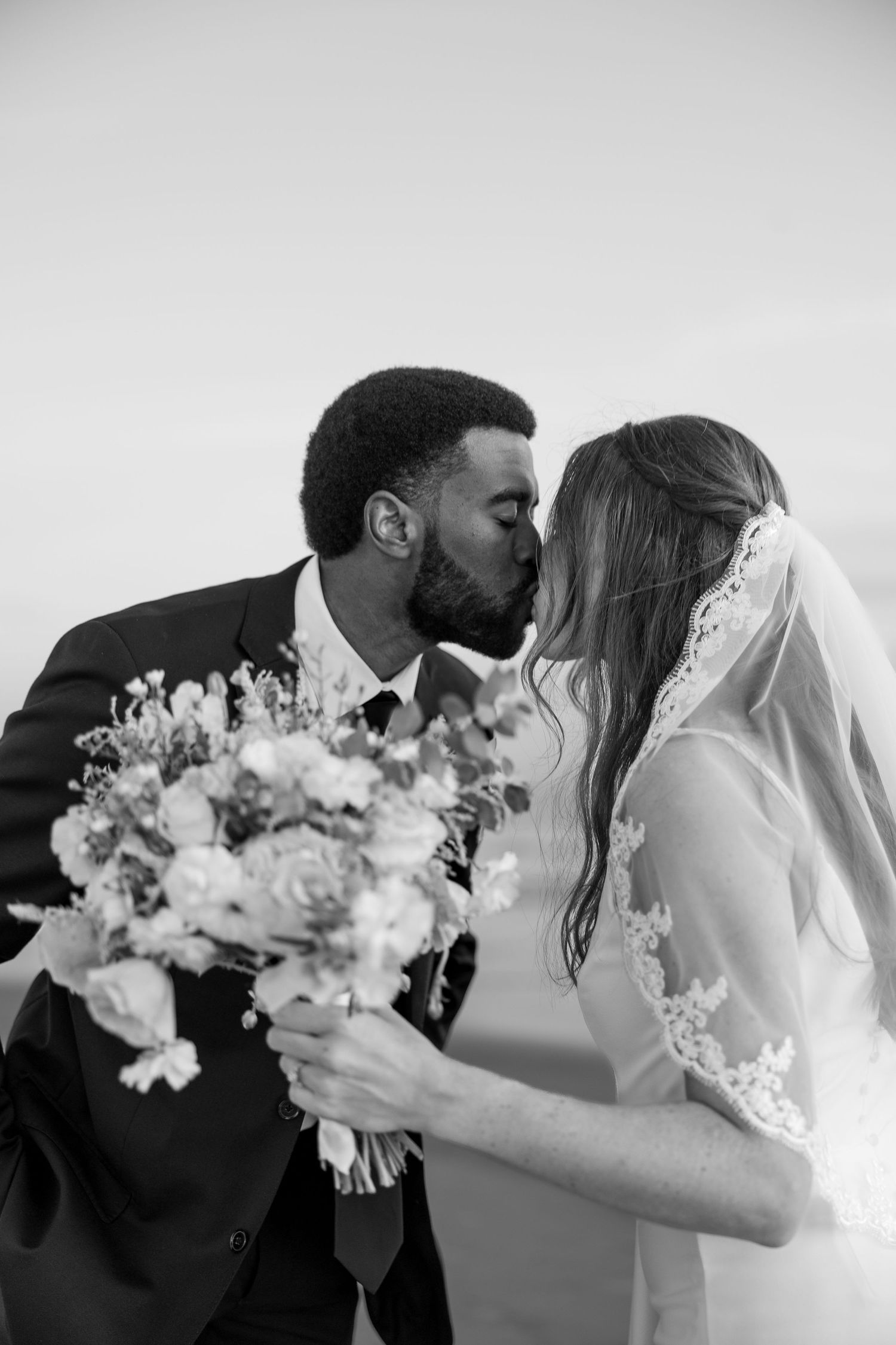 Black and white photo of an interracial couple kissing on their wedding day, bride holding a floral bouquet.