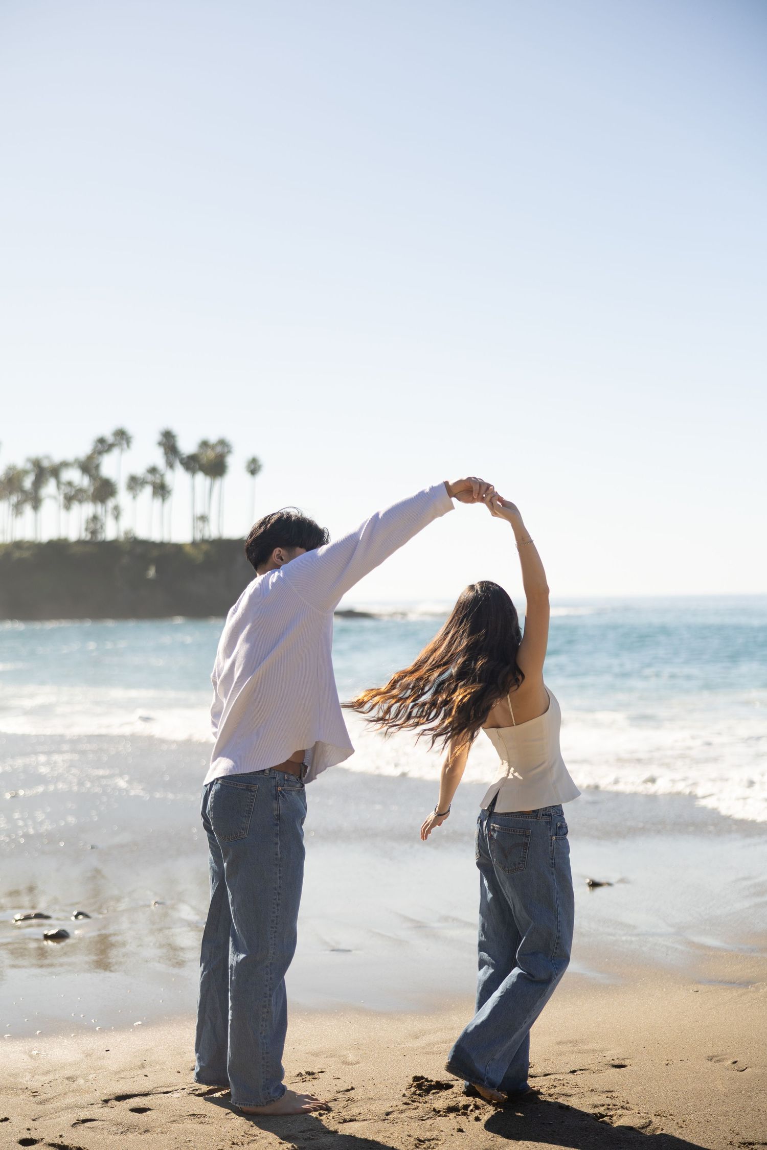 Couple dancing on a sunny beach with ocean waves and palm trees in the background, wearing jeans and white tops.