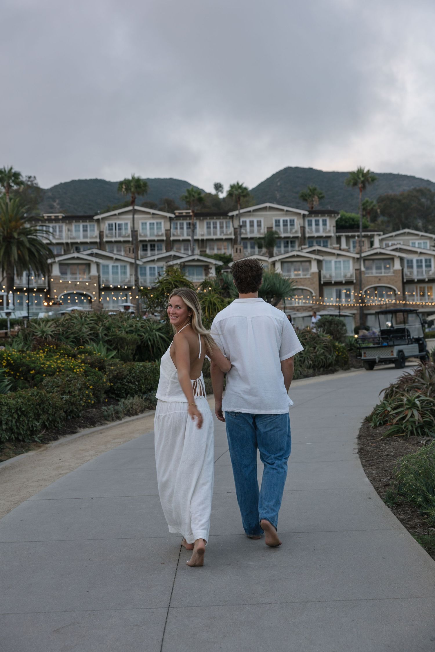 Couple walking hand in hand on a curved path toward a coastal resort with palm trees and hills at dusk.