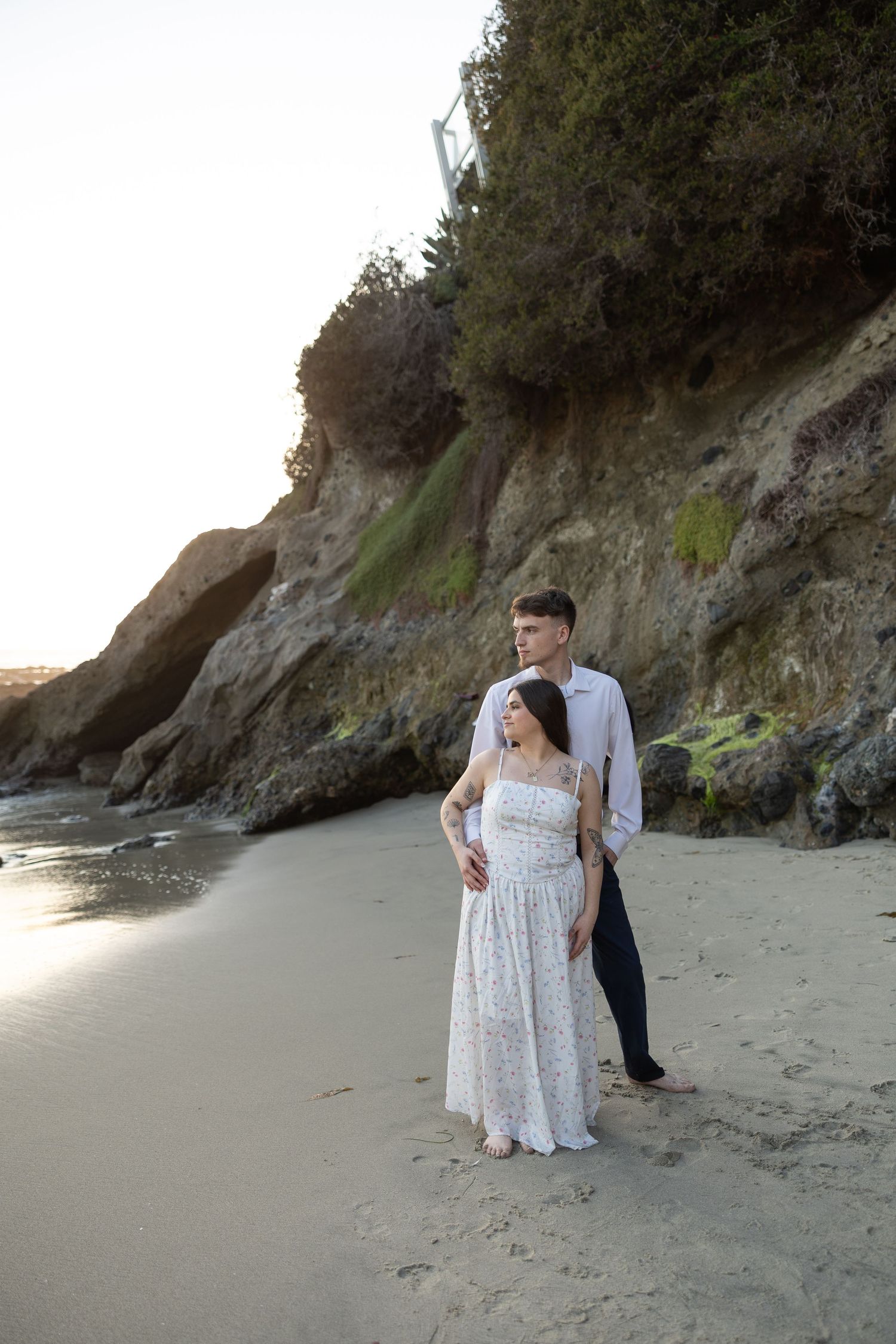 A couple takes romantic photos on a sandy beach with cliffs in the background.
