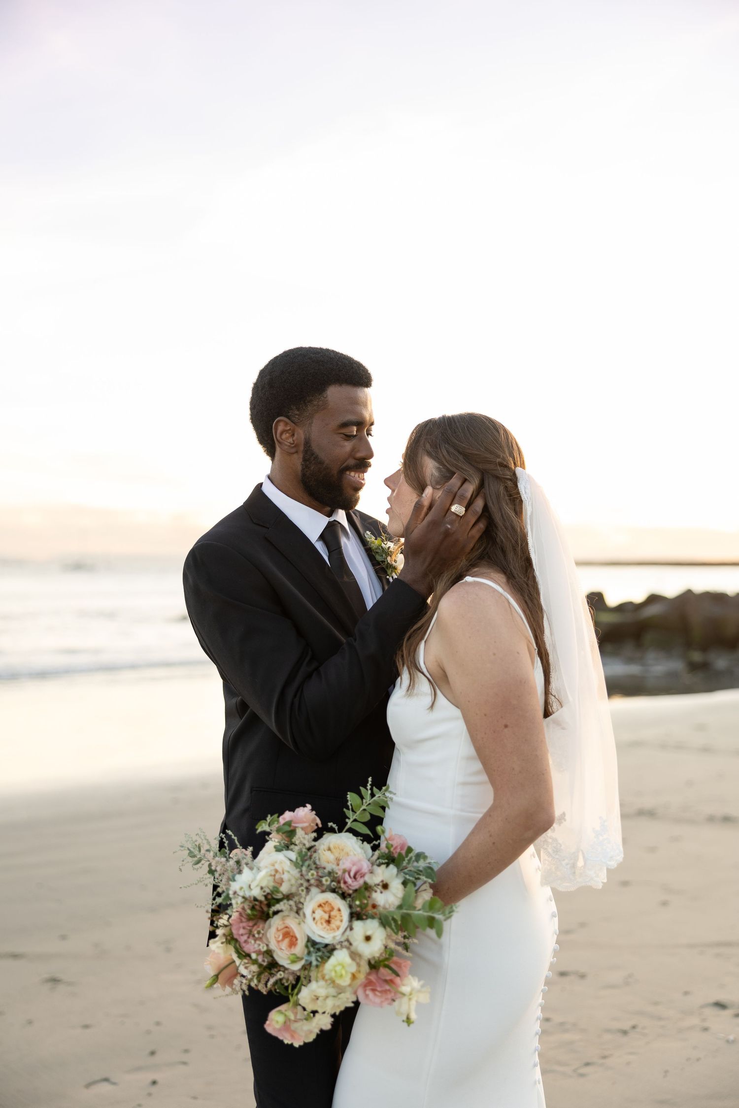 Bride and groom share a tender moment on the beach at sunset, with bride holding a pastel floral bouquet.
