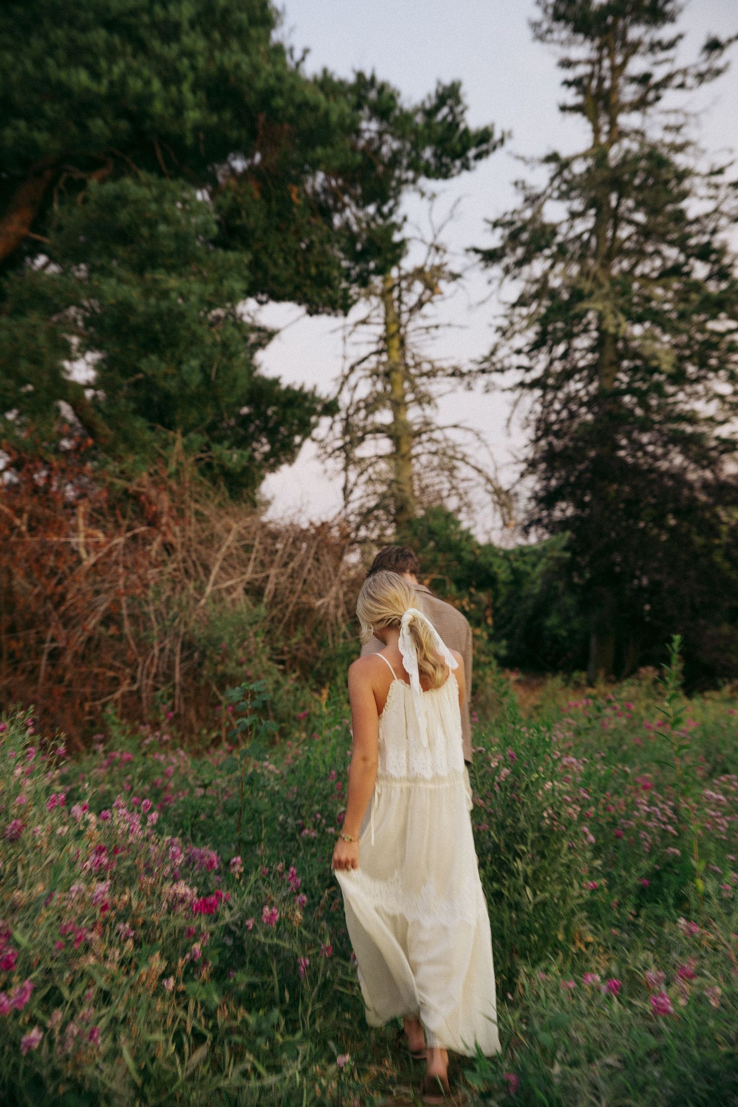 A woman in a flowing white maxi dress walks through a wildflower meadow surrounded by lush green trees.