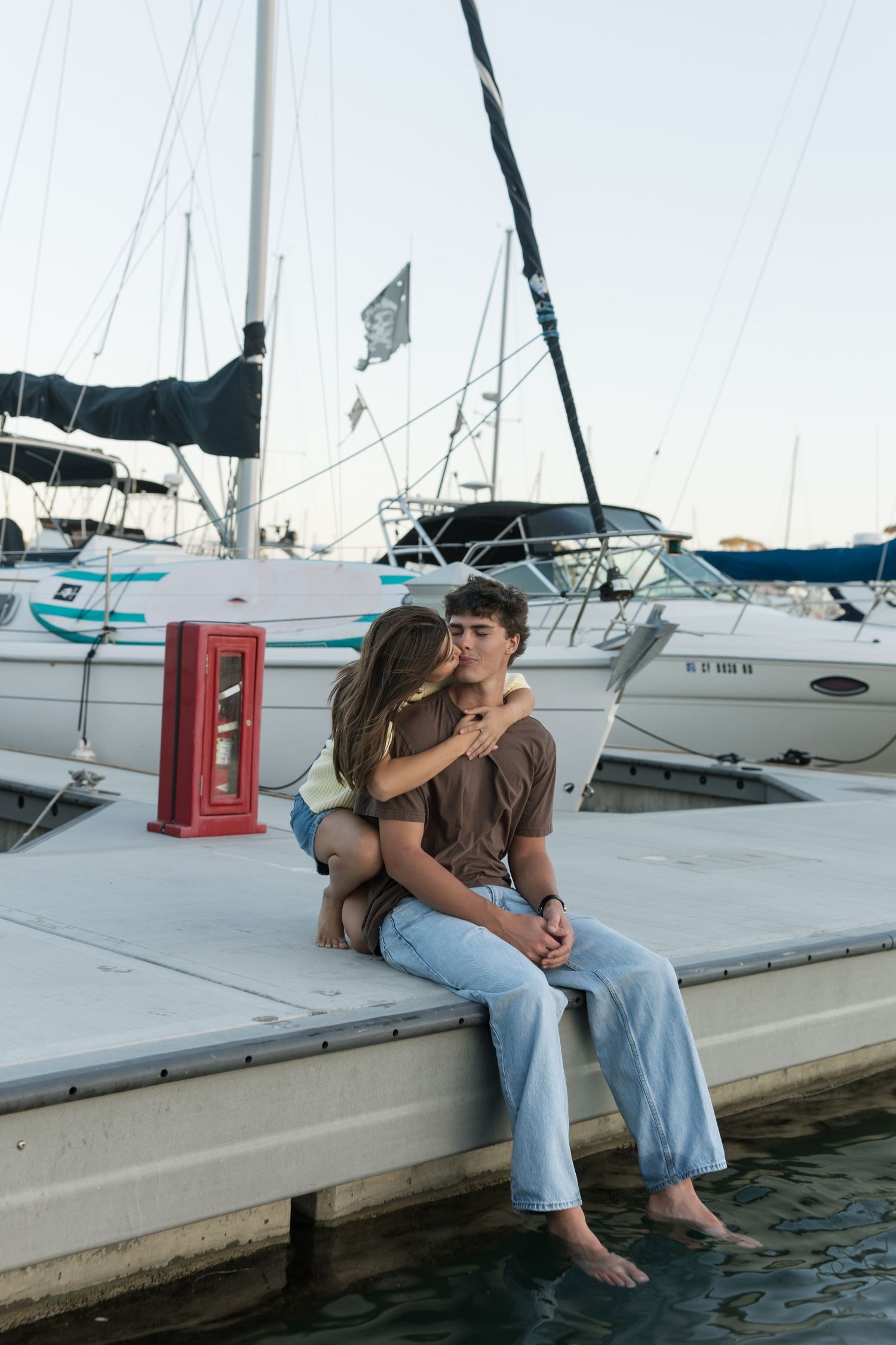 Couple sitting barefoot and embracing on a marina dock beside docked sailboats and a red box, with calm water below.