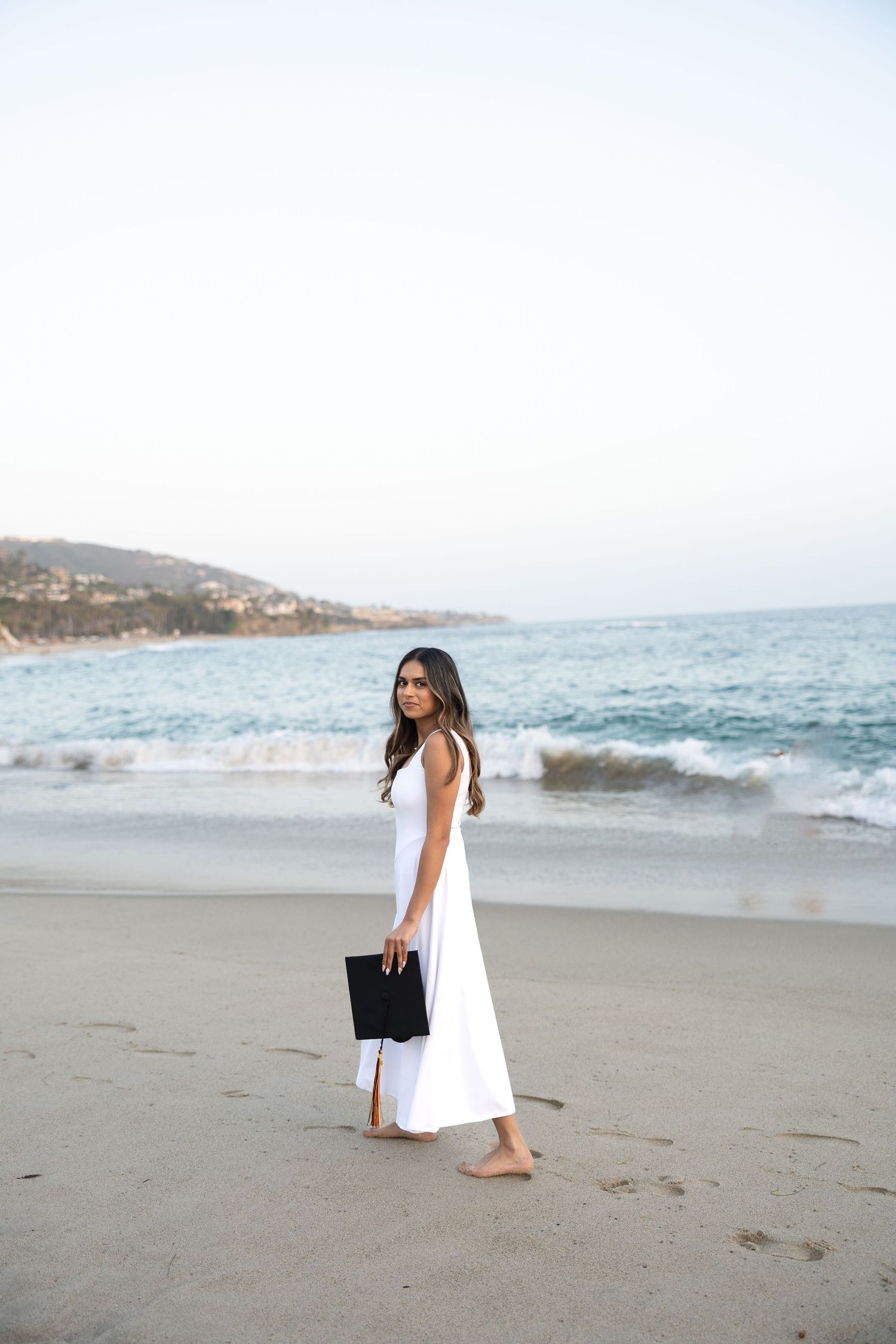 Woman in white maxi dress holding black clutch, walking barefoot on sandy beach with ocean waves in background.