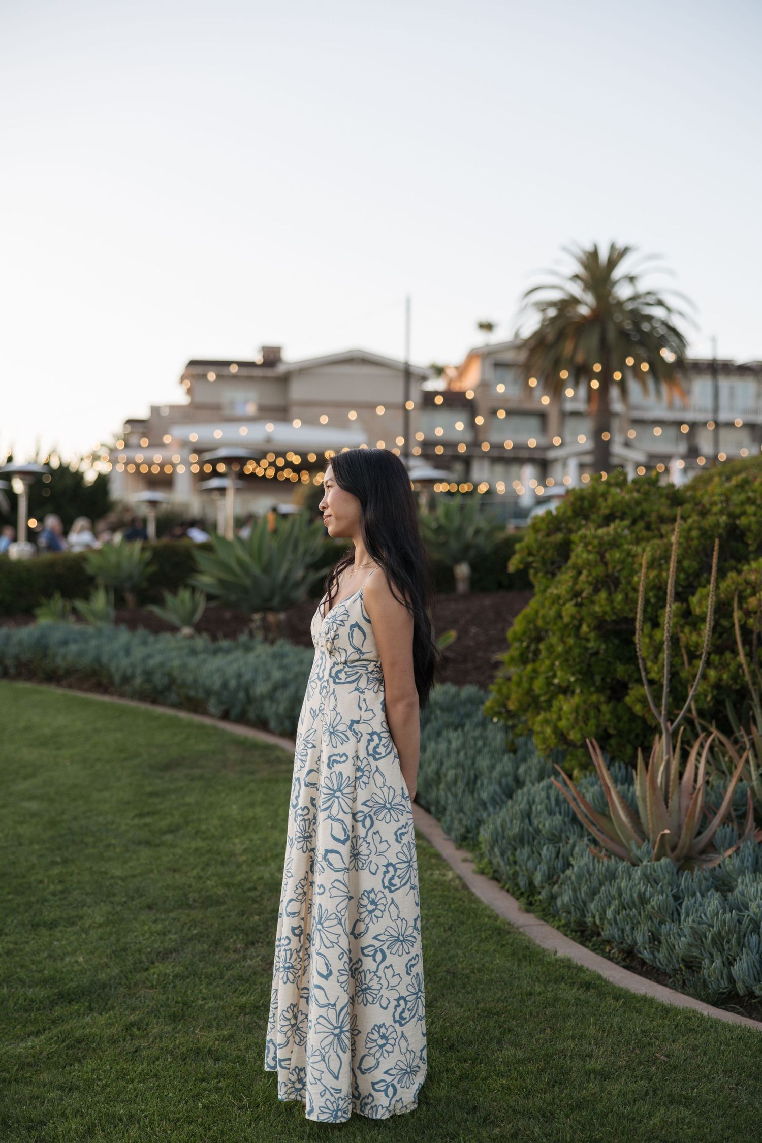 Woman in floral maxi dress standing on green lawn at dusk with string lights and palm trees in background.