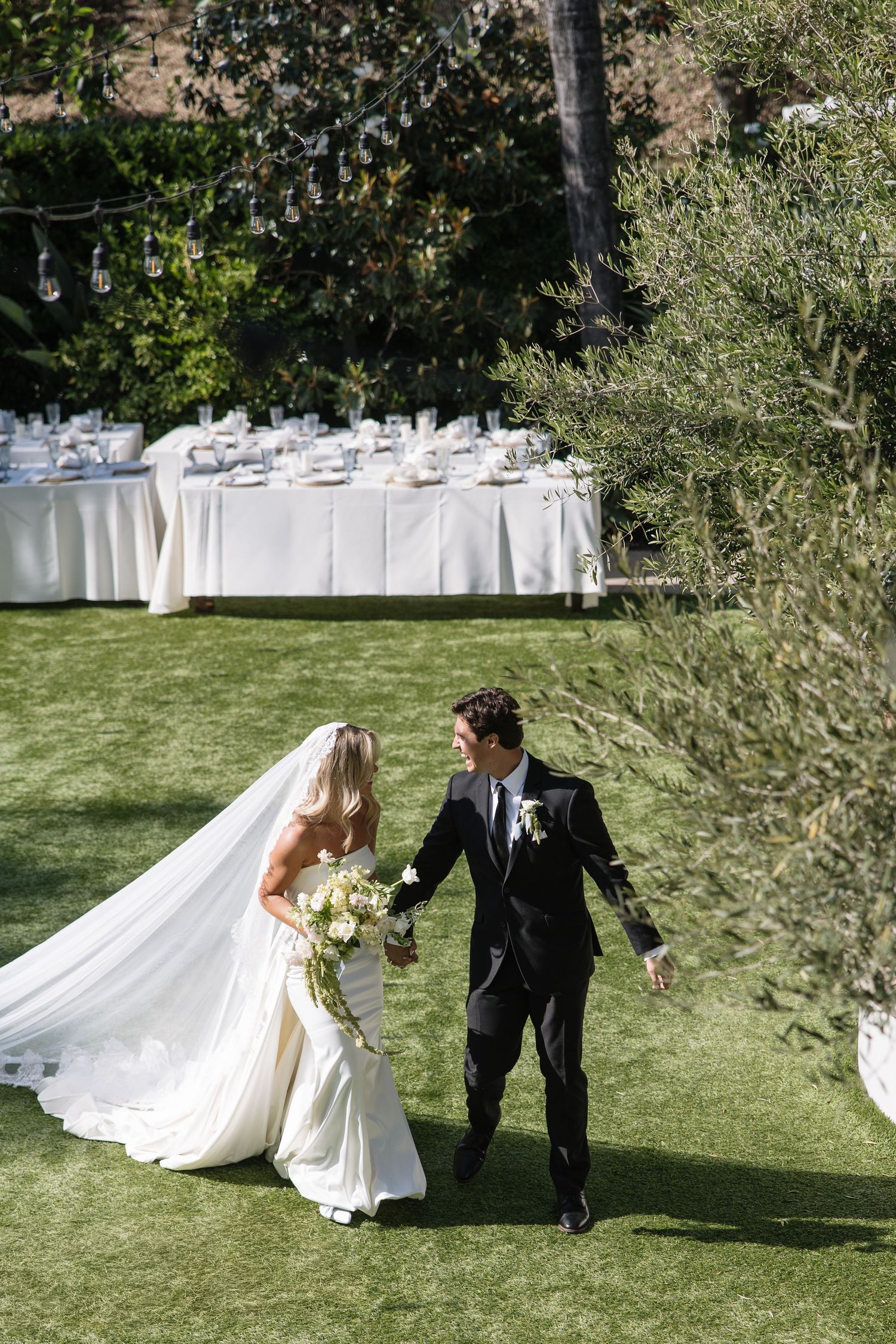Bride and groom walk hand-in-hand on lush green lawn with elegant outdoor reception tables in background.
